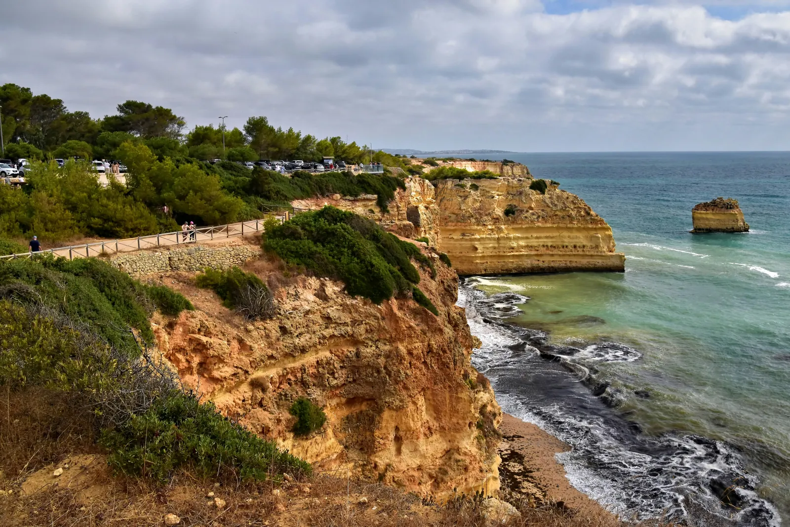 Golden sandstone cliffs covered in green scrub dropping to a rocky shore and turquoise sea, with a fenced path along the clifftop, pine trees behind and a single rock stack offshore under a cloudy sky