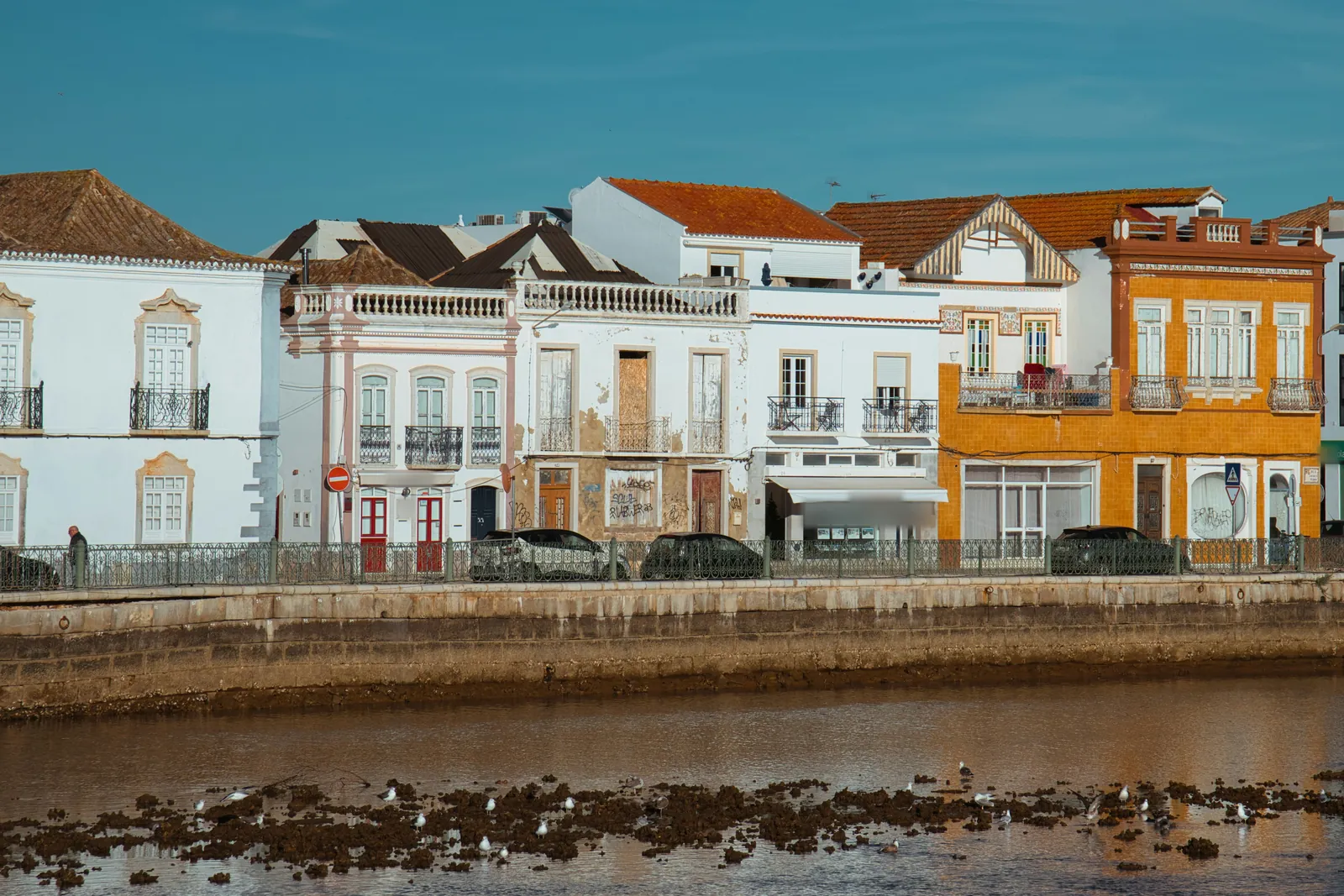 A row of old two-storey town houses, white, cream and orange-tiled, with wrought-iron balconies and tiled roofs, above a stone waterfront wall, with gulls on the mud and weed of the water at low tide under a deep blue sky