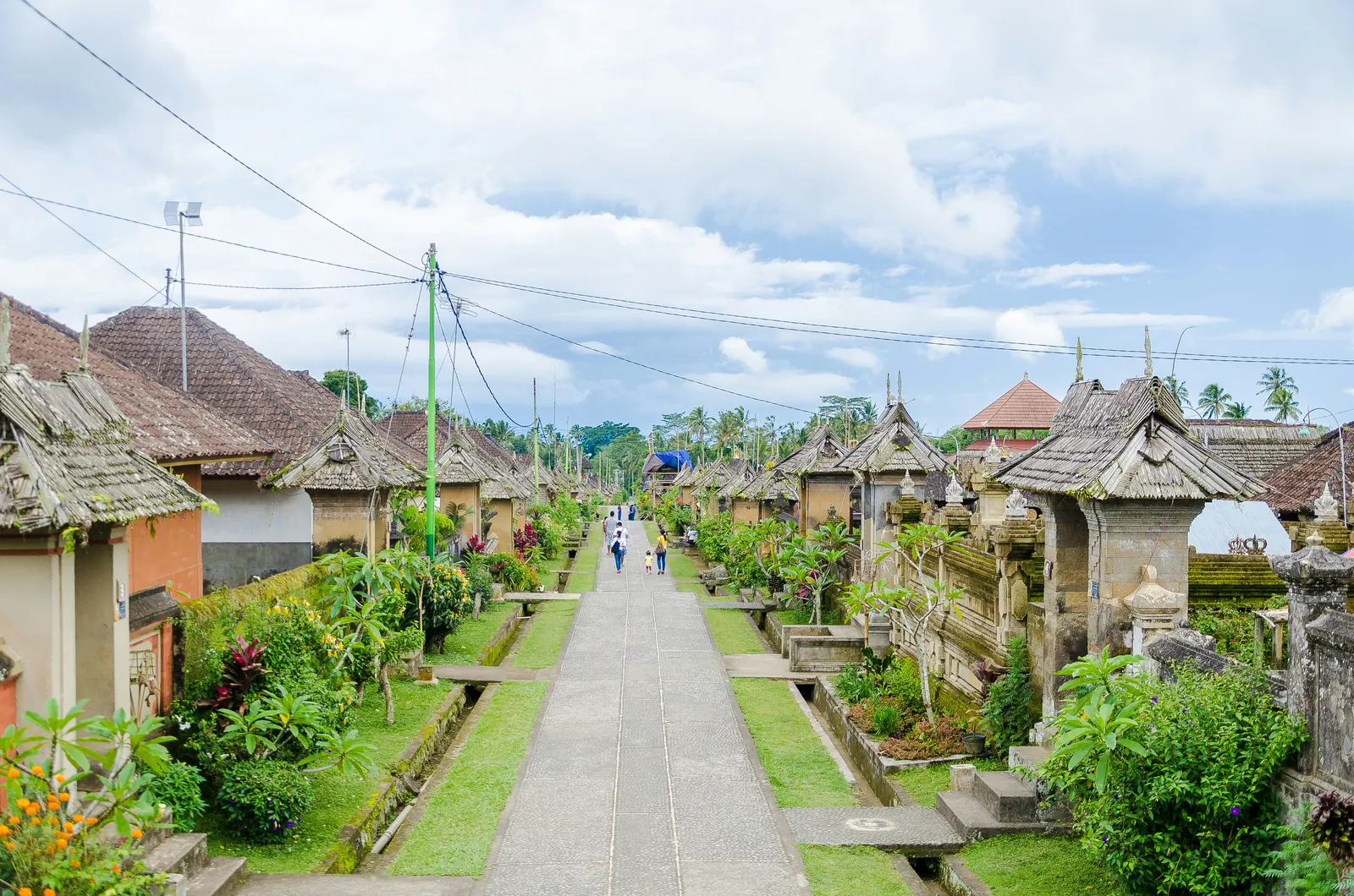 Long paved street in Penglipuran village lined with gardens, stone gateways and traditional bamboo-roofed houses