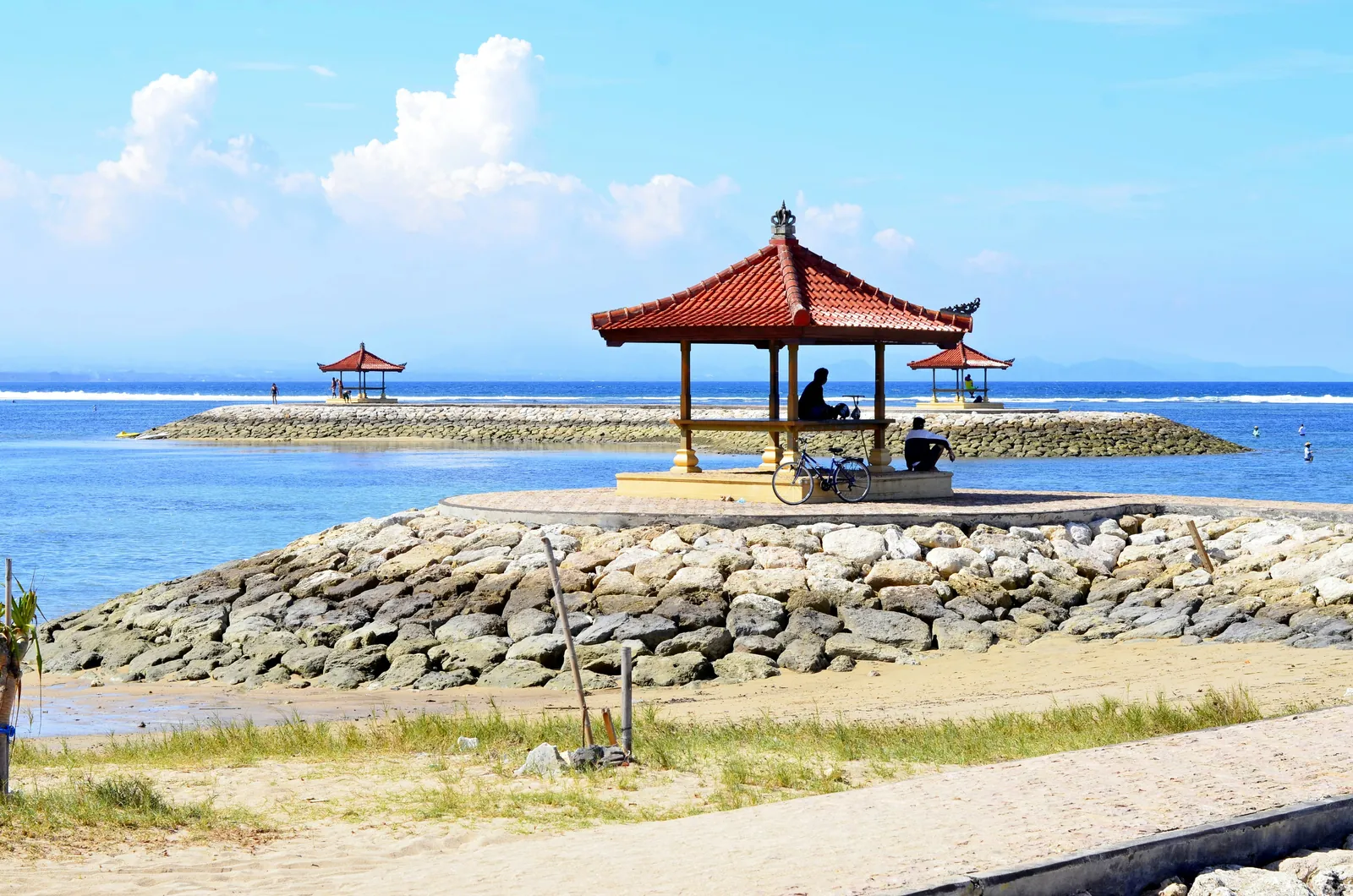 Red-roofed pavilion on a stone breakwater at Sanur Beach, with a sandy path in front and calm blue water behind