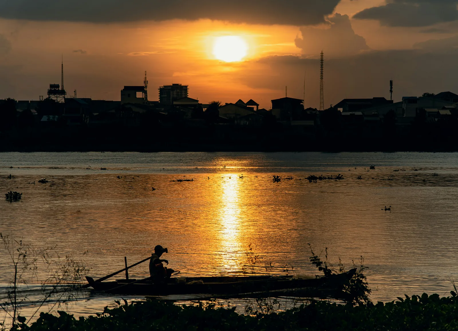 Fishing boat in silhouette on a golden river at sunset, with low rooftops and towers on the far bank in Phnom Penh
