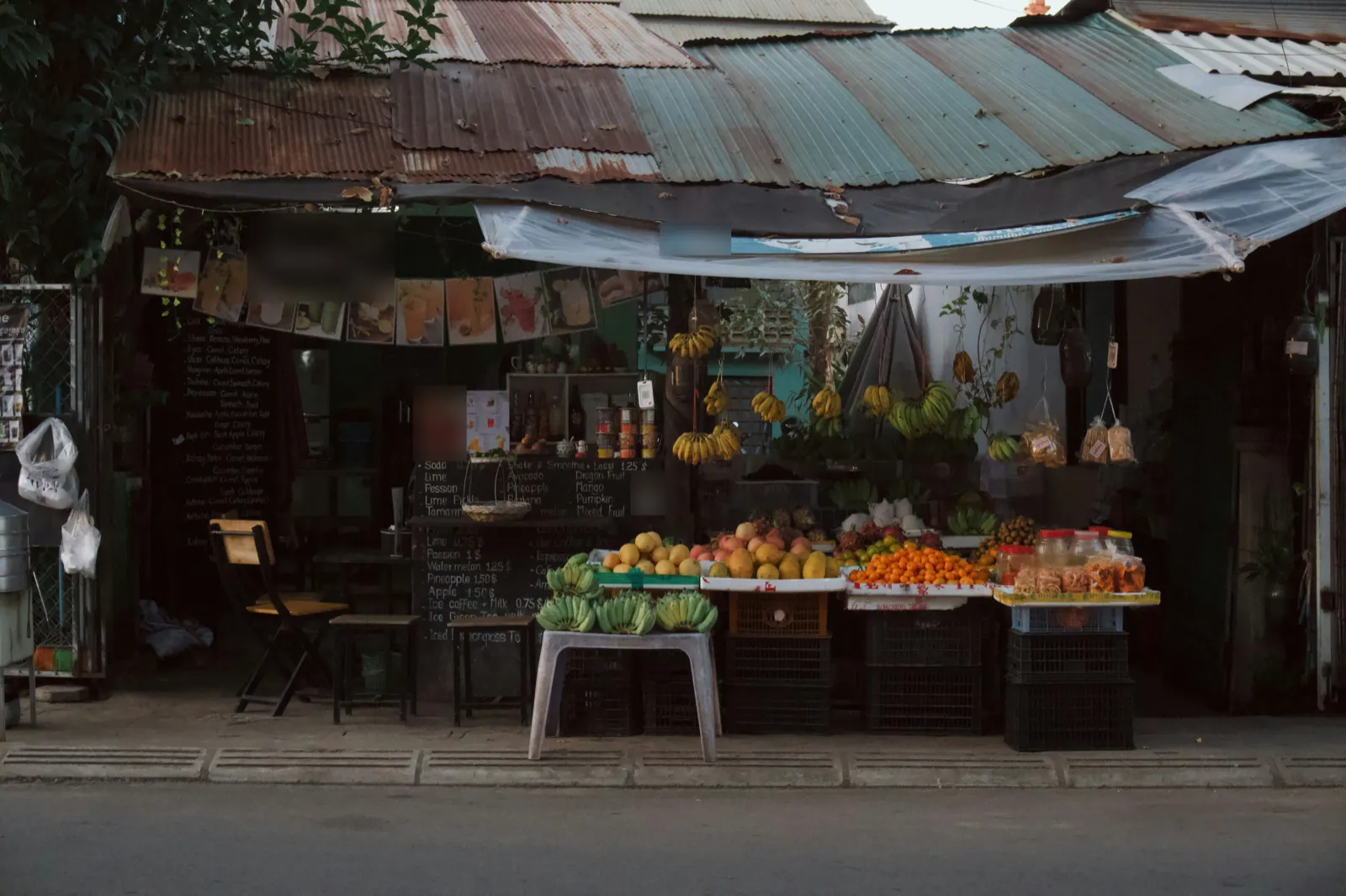 A roadside fruit stall under a rusty iron roof in Siem Reap, with bunches of bananas hanging and mangoes, apples and mandarins piled on crates