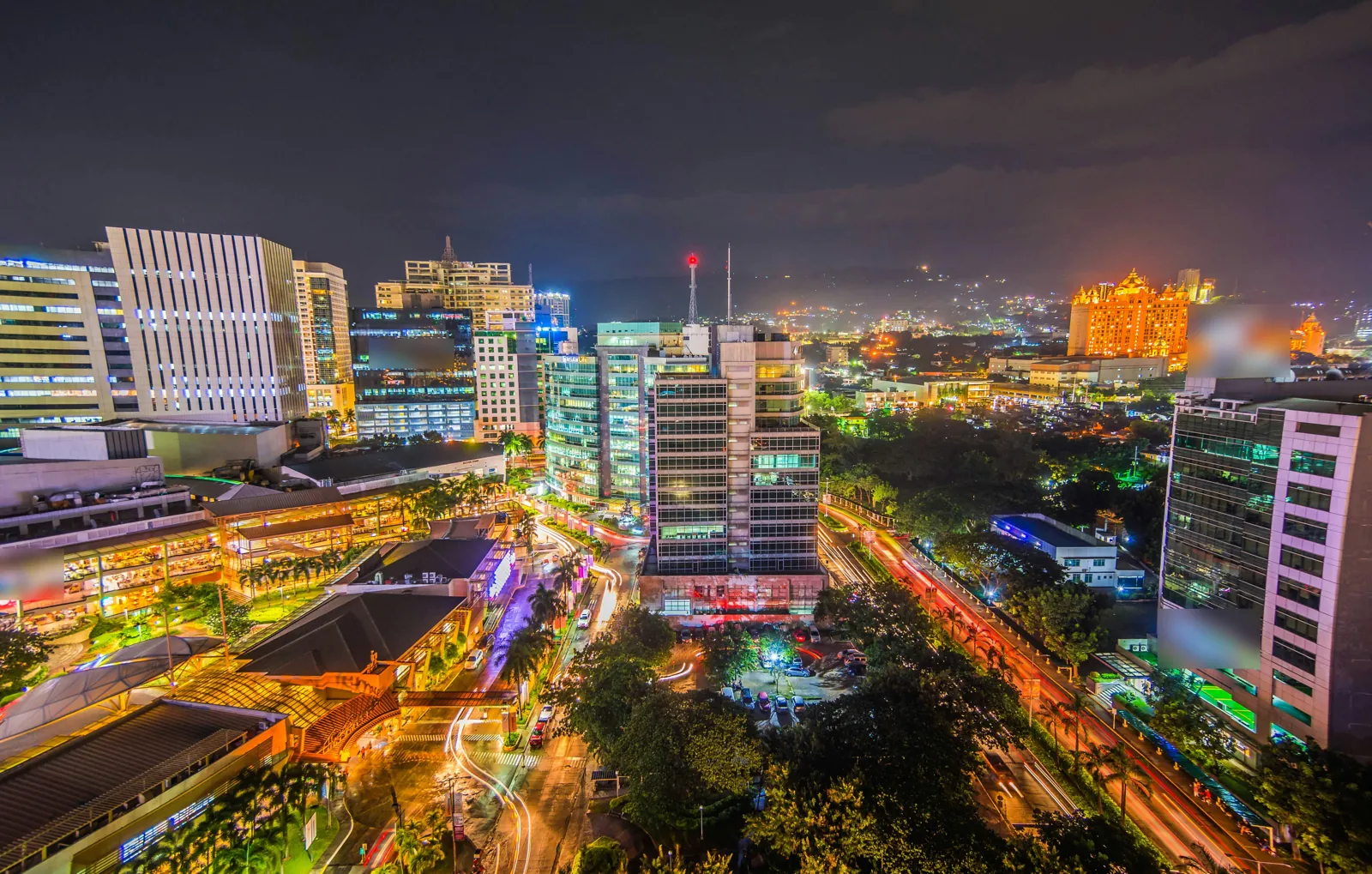 Office towers, a lit shopping centre and roads streaked with car lights at night, with hills and the lights of houses behind