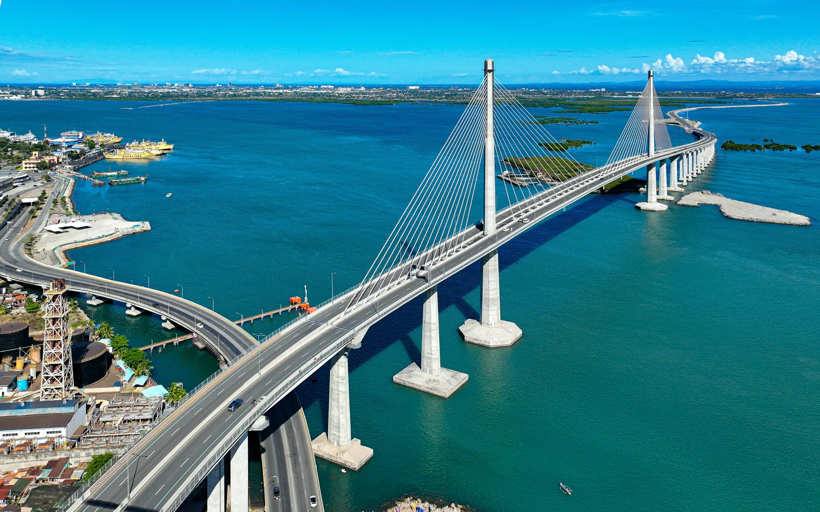 A cable-stayed bridge with two tall towers carries a road across blue water, with a port and the city on the far shore