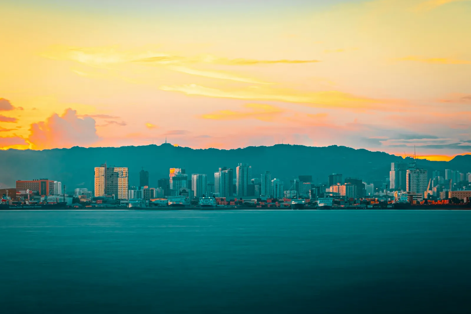 A city skyline of towers below green hills, seen across calm water under an orange and pink sunset sky