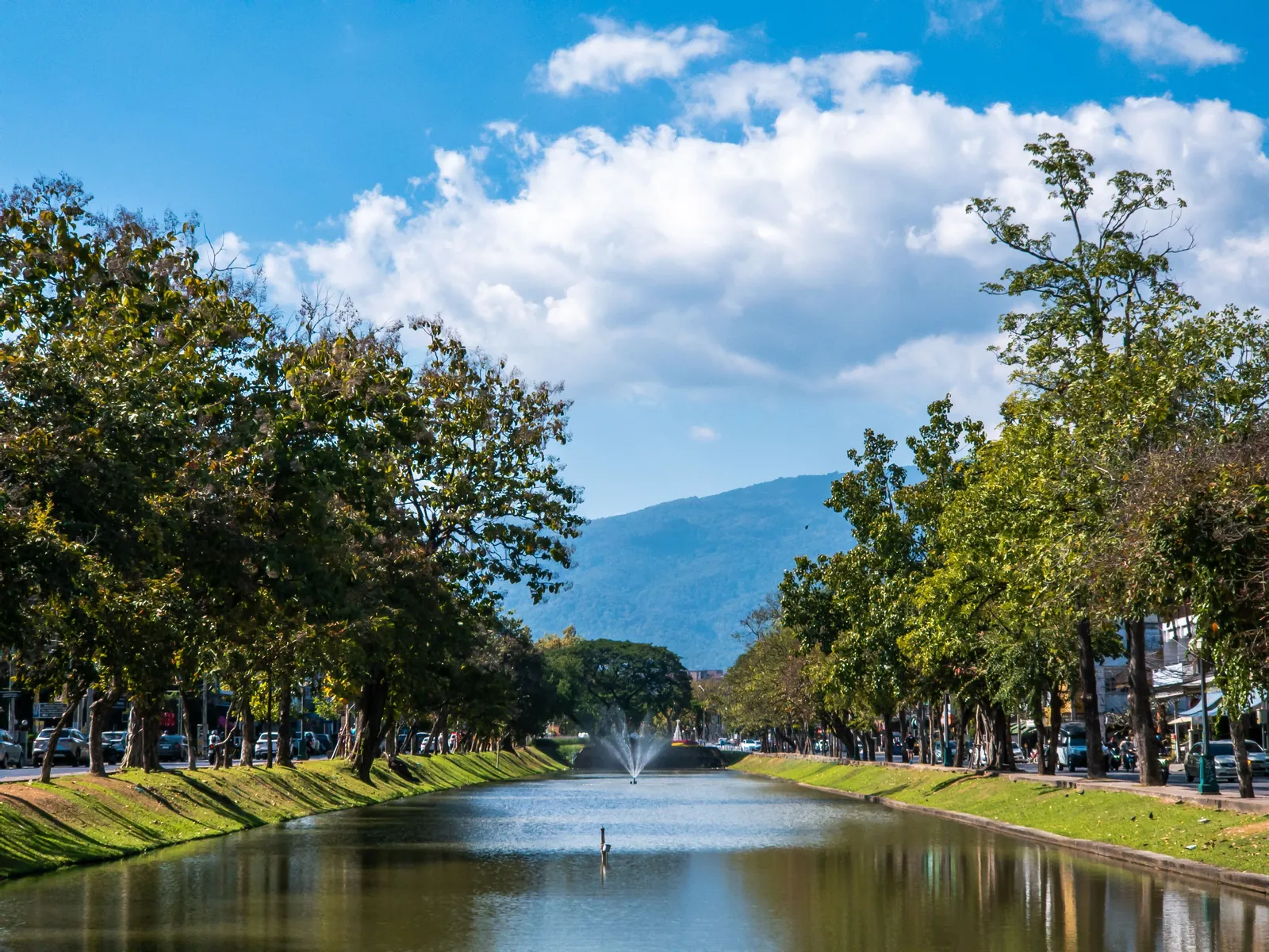 A straight canal with grassy banks and a fountain in the middle, lined on both sides by tall leafy trees, roads and parked cars, with a forested mountain behind under a blue sky and white clouds