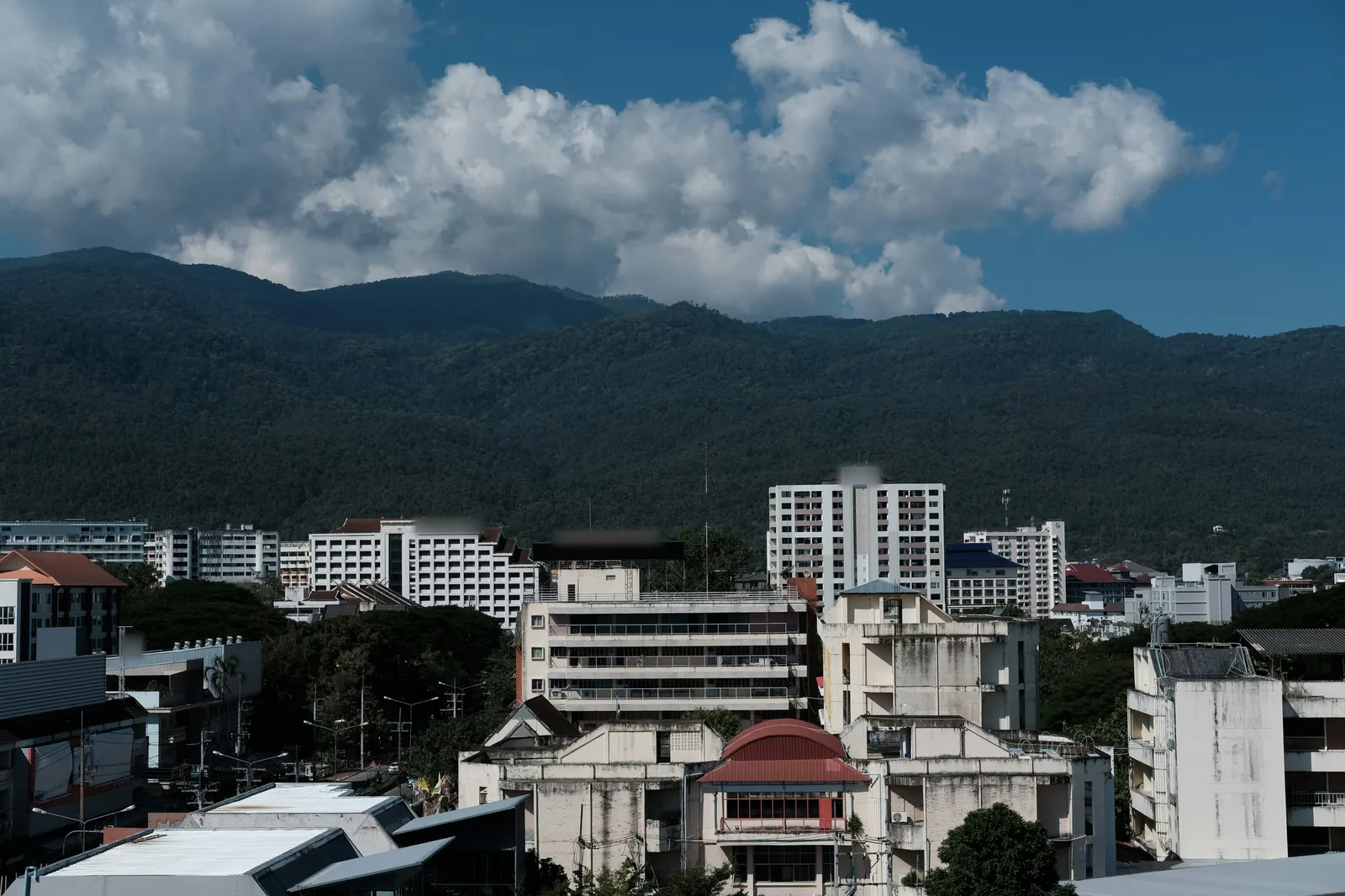 Low-rise and mid-rise concrete buildings and apartment blocks in the foreground, with a long, dark green forested mountain ridge behind under a blue sky and large white clouds