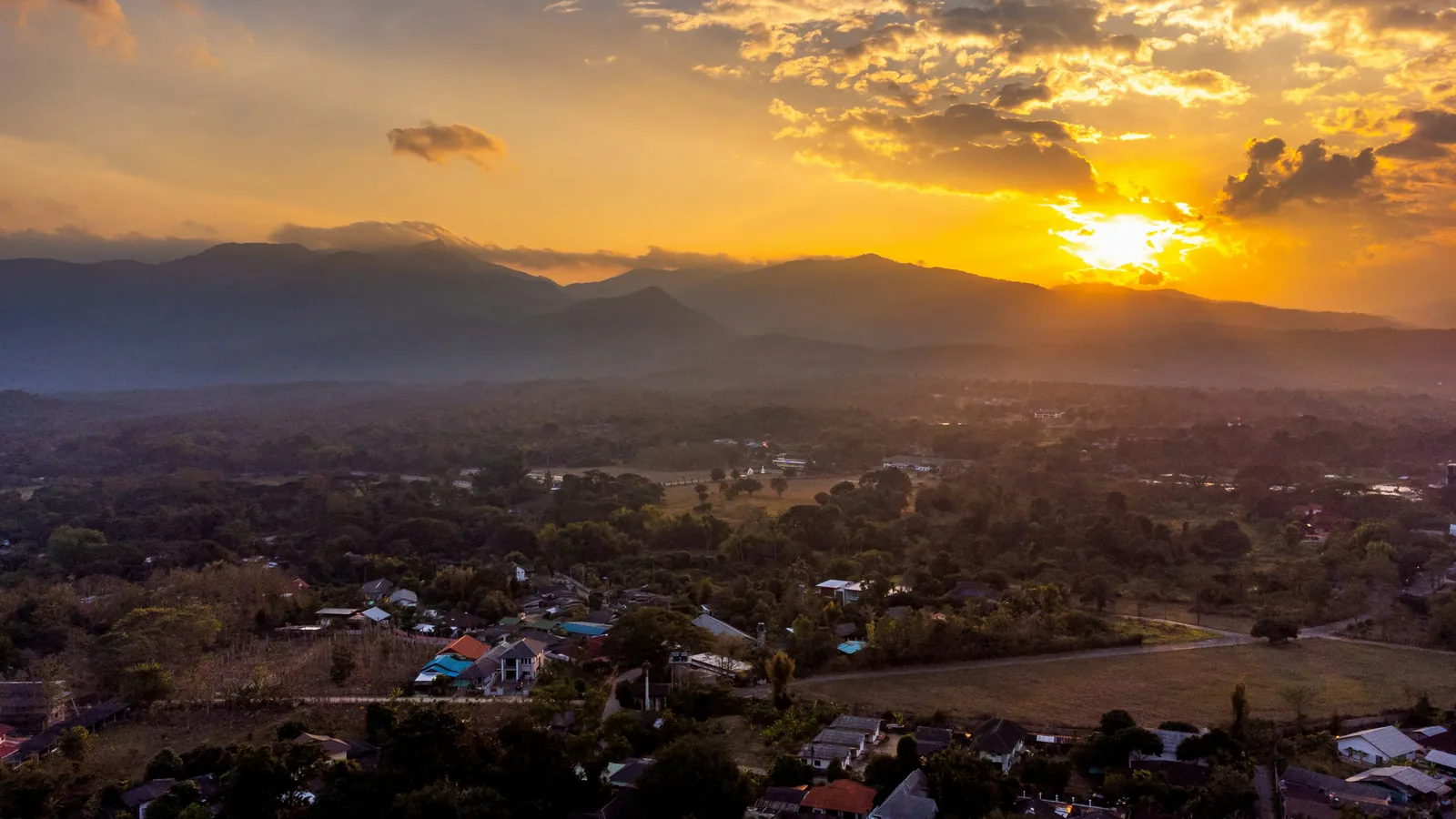 Houses with coloured roofs scattered among trees and dry fields in a wide valley, seen from the air, with layered mountain ridges behind and the sun setting in a golden sky
