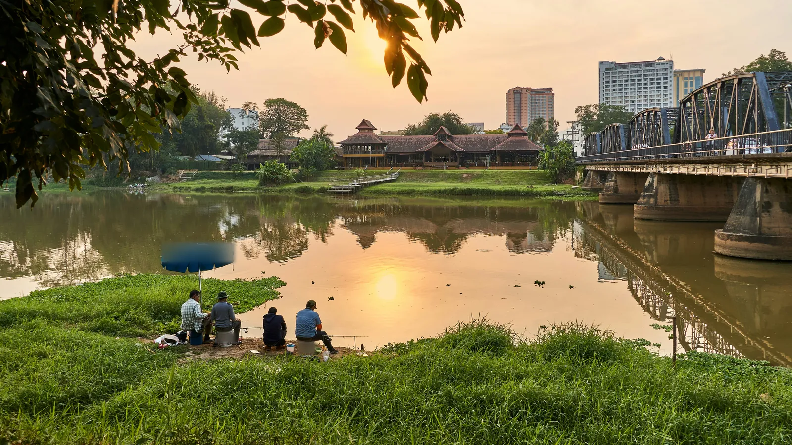 Four men sitting on stools and buckets on a grassy riverbank at sunset, fishing rods out, one under a blue umbrella, with the sun reflected in the brown water, an old steel bridge on the right and a long timber building across the river