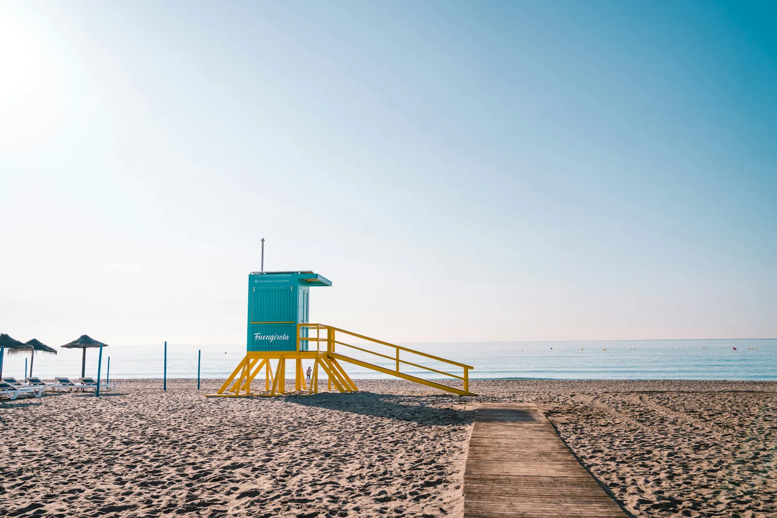 A blue and yellow lifeguard tower on an empty sandy beach, with sun umbrellas to one side and a calm sea in the morning light