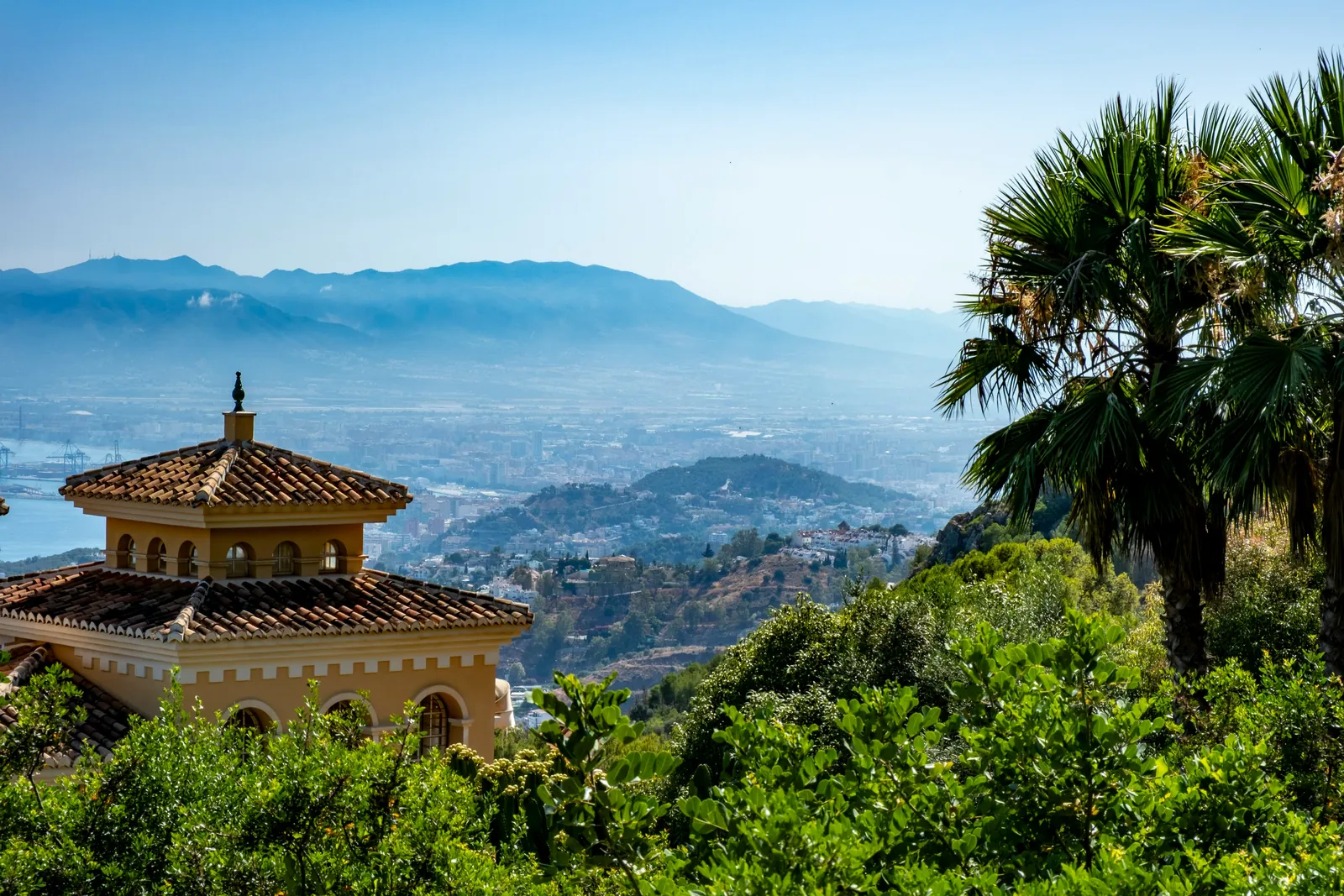 A tiled-roof pavilion among palm trees on a green hillside, looking out over a city spread across the plain to blue mountains