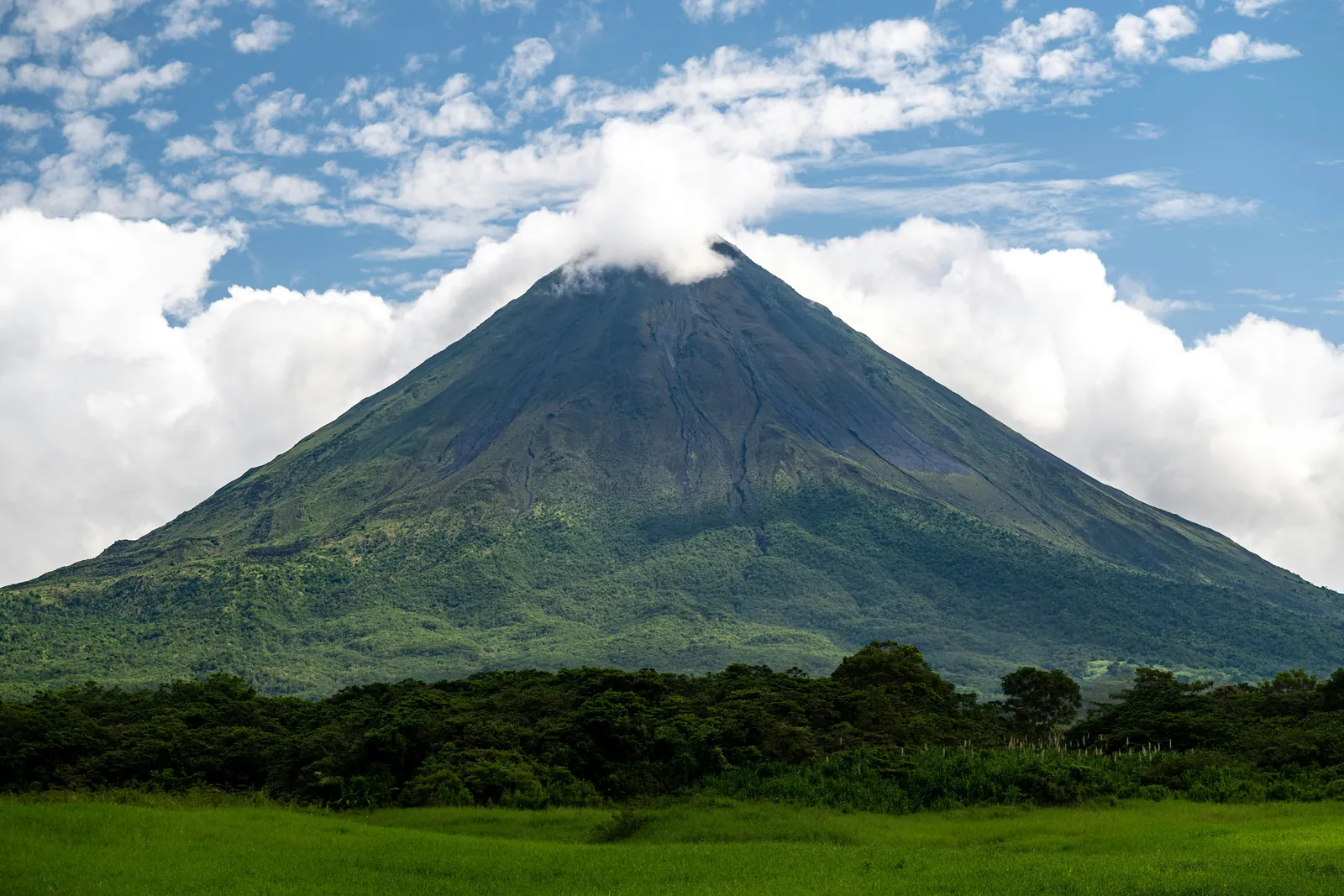 A symmetrical green volcano cone with a small cloud on its summit, rising above dense forest and a bright green field under a blue sky with white clouds
