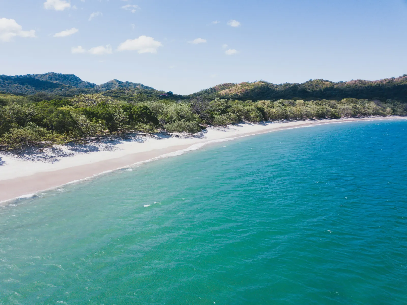A long curving white sand beach lined with dry forest, turquoise water lapping the shore and green hills behind, seen from a drone