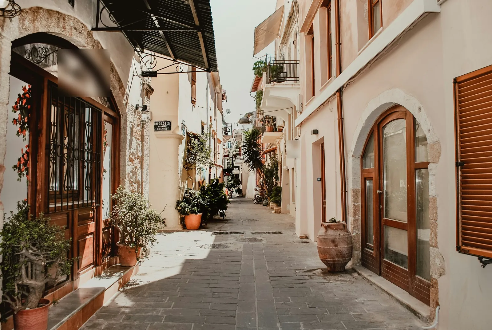 A narrow paved lane between cream houses with arched wooden doors, a large clay pot and potted plants along both sides