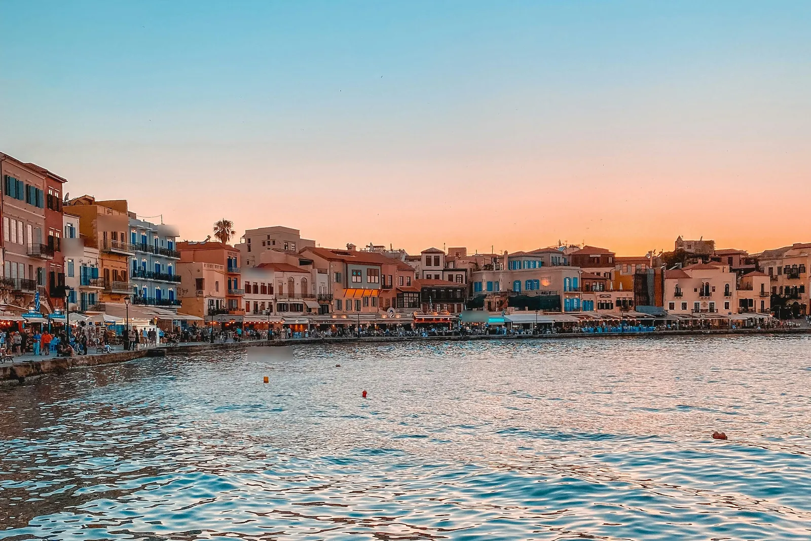 Calm blue water in front of a long row of pastel three and four storey buildings with balconies and blue shutters, cafés along the quay, under an orange sunset sky