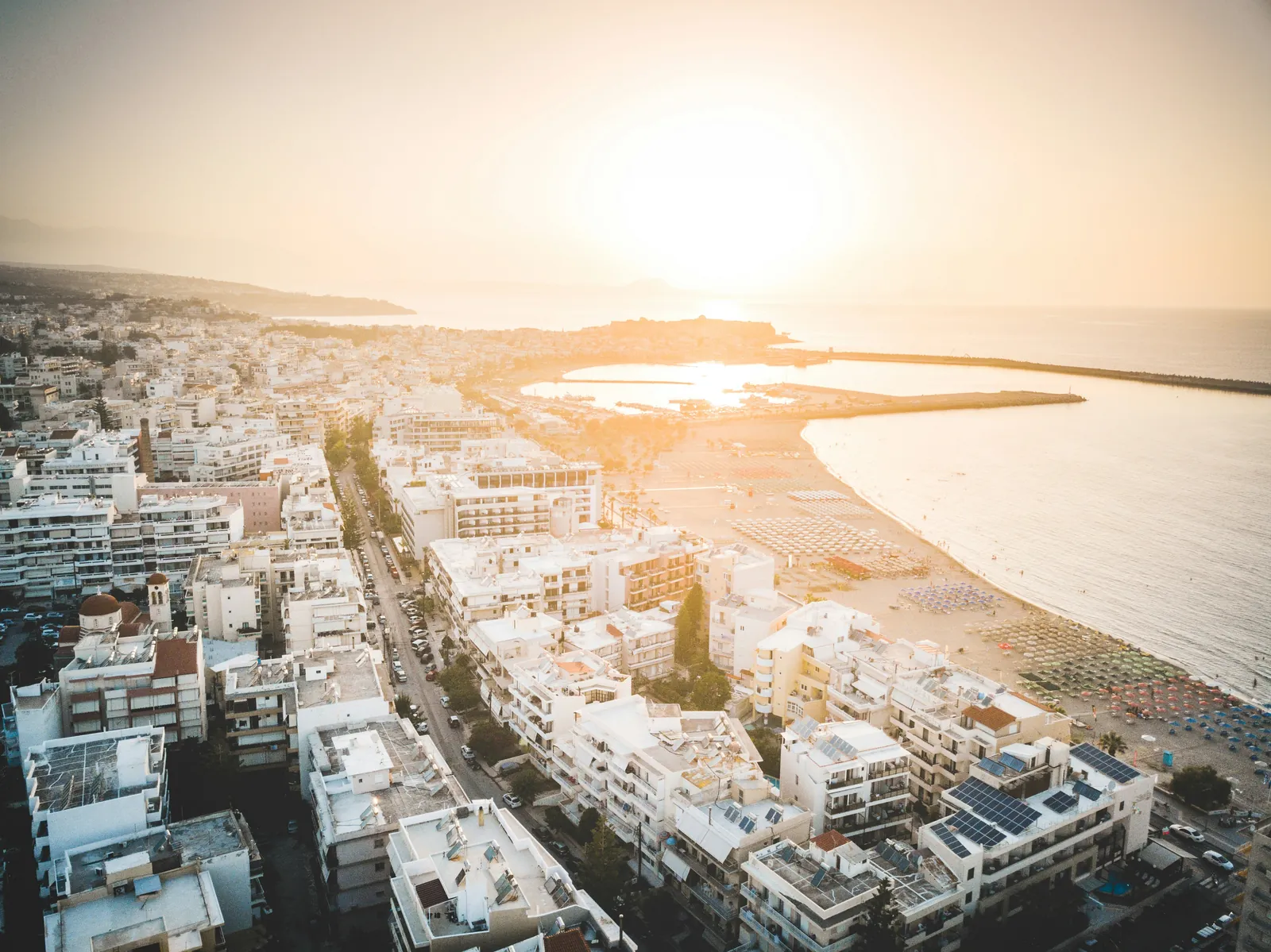 White apartment blocks with solar panels on their roofs above a long sandy beach lined with umbrellas, with a harbour wall and the sea behind, seen from the air
