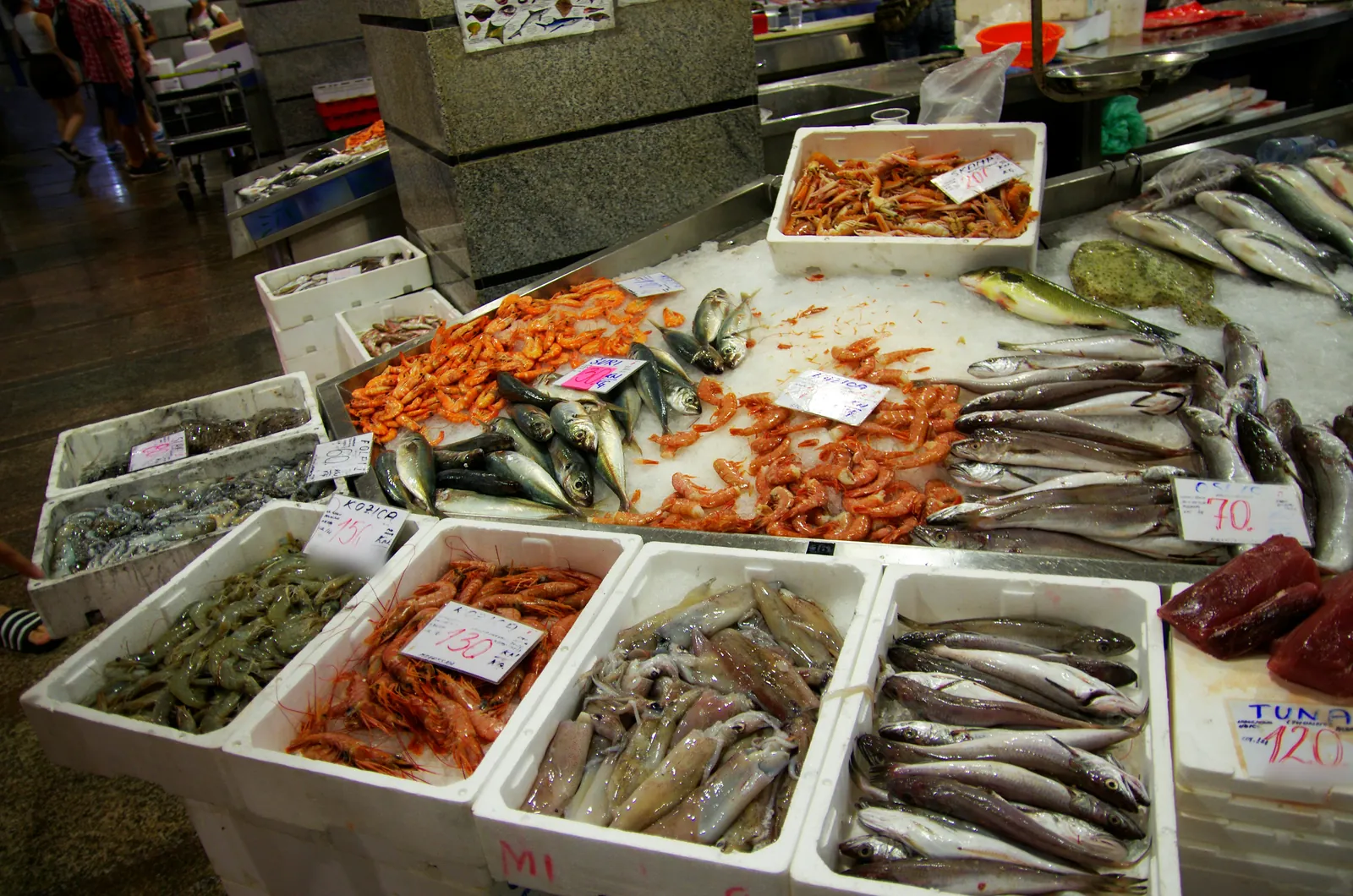 Fish market counter with boxes of prawns, squid, hake and mackerel on ice, and handwritten price cards in kuna per kilo
