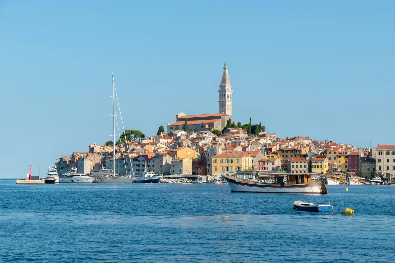 Rovinj old town of colourful houses crowding a small hill topped by a church and tall bell tower, with yachts and a wooden boat on calm blue water in front