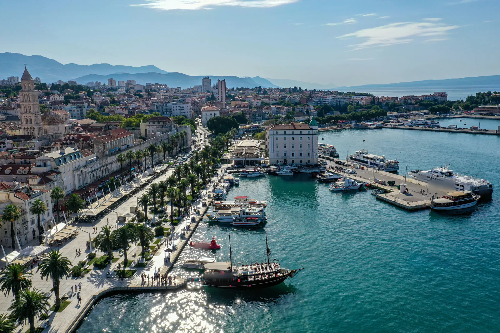 Aerial view of Split’s palm-lined waterfront promenade beside the old palace walls and a bell tower, with tour boats, ferries and a wooden sailing ship in the turquoise harbour