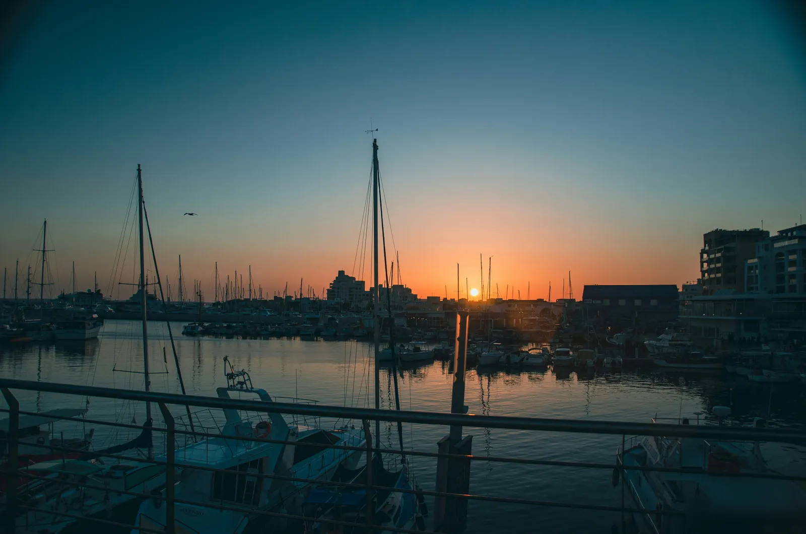 Fishing boats and yacht masts silhouetted in Limassol harbour at sunset, with the sun low behind the waterfront