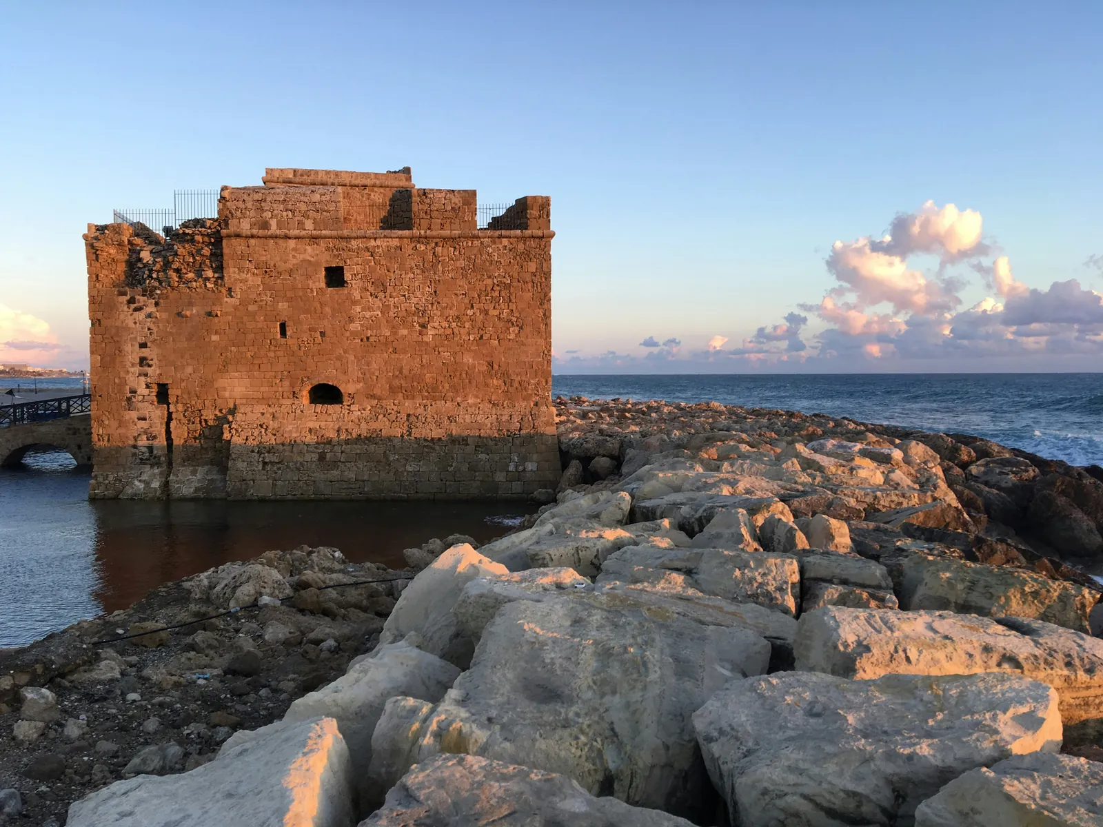 The square stone fort at the end of Paphos harbour glowing orange at sunset, with breakwater rocks in front and the sea behind