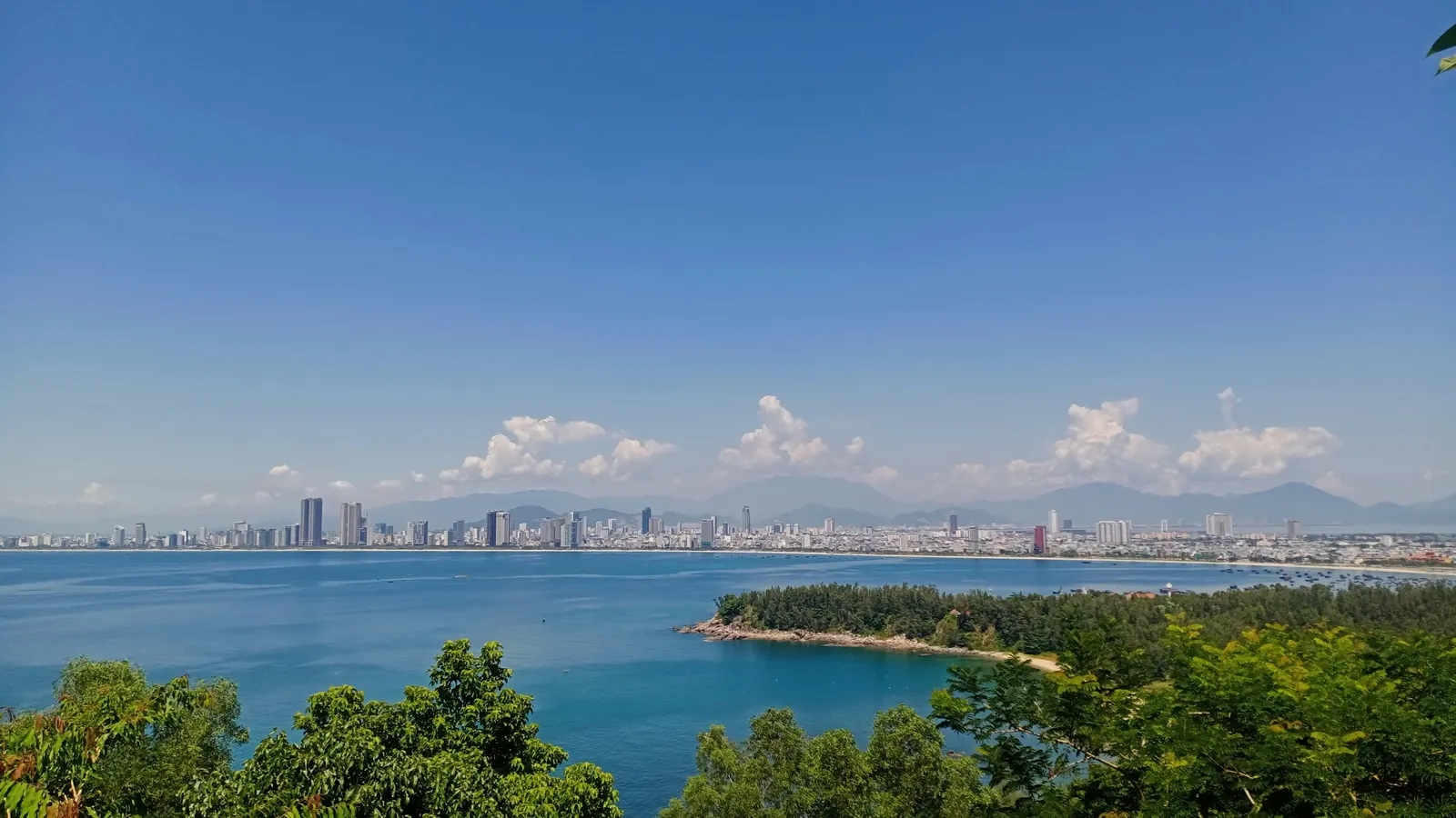 A wide blue bay seen from a wooded hillside, with a low forested spit reaching into the water and a long line of city buildings on the far shore below hazy mountains and small white clouds