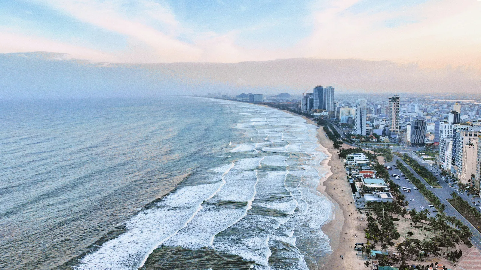 A long curving sandy beach with lines of white surf rolling in, seen from the air, with a park of palm trees, a seafront road and rows of high-rise buildings on the right, under a pastel evening sky