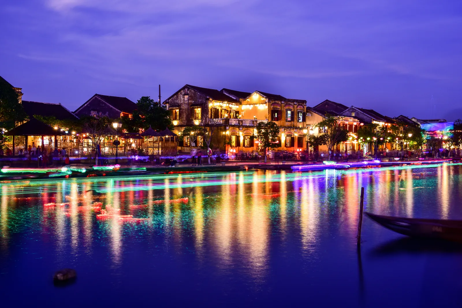 A row of old two-storey houses with tiled roofs and lit windows along a riverbank at dusk under a deep blue sky, with streaks of coloured light from moving boats reflected in the water and the bow of a wooden boat at the lower right