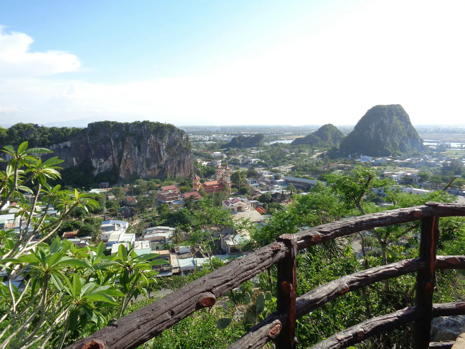 Steep grey and green limestone outcrops rising from a flat plain of houses and trees, seen past a rustic wooden-look railing and frangipani leaves, under a pale blue sky