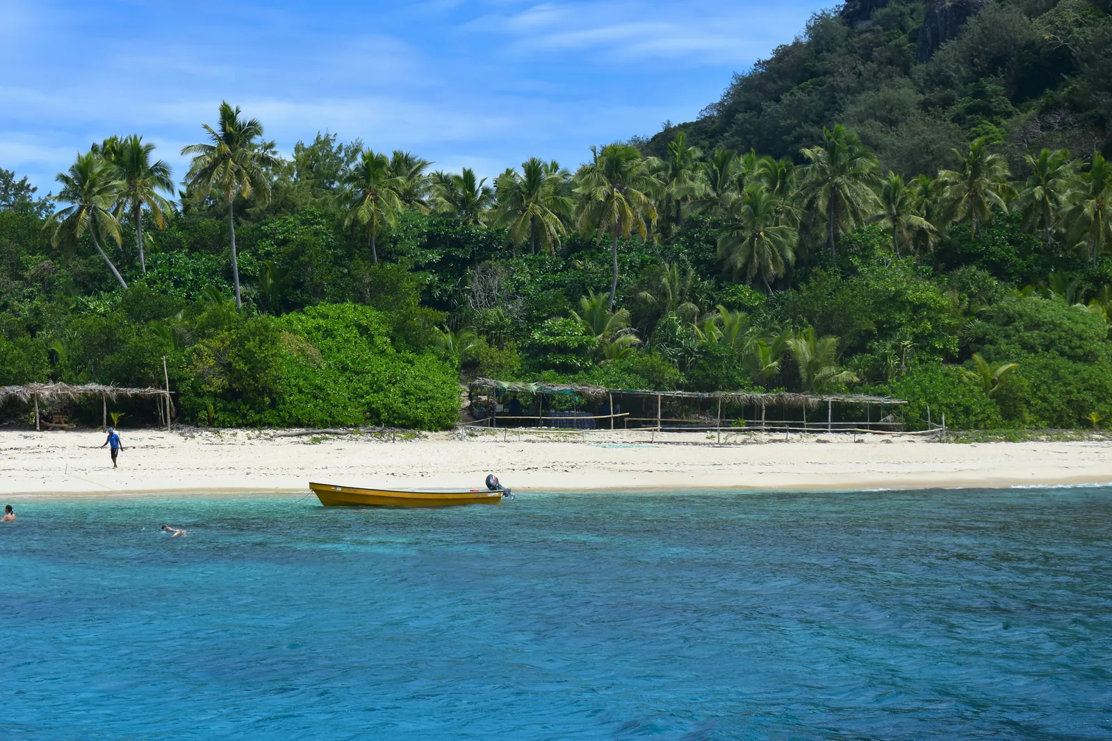 A yellow boat anchored off a white sand beach backed by coconut palms and thick green forest on Monuriki island, seen from the water