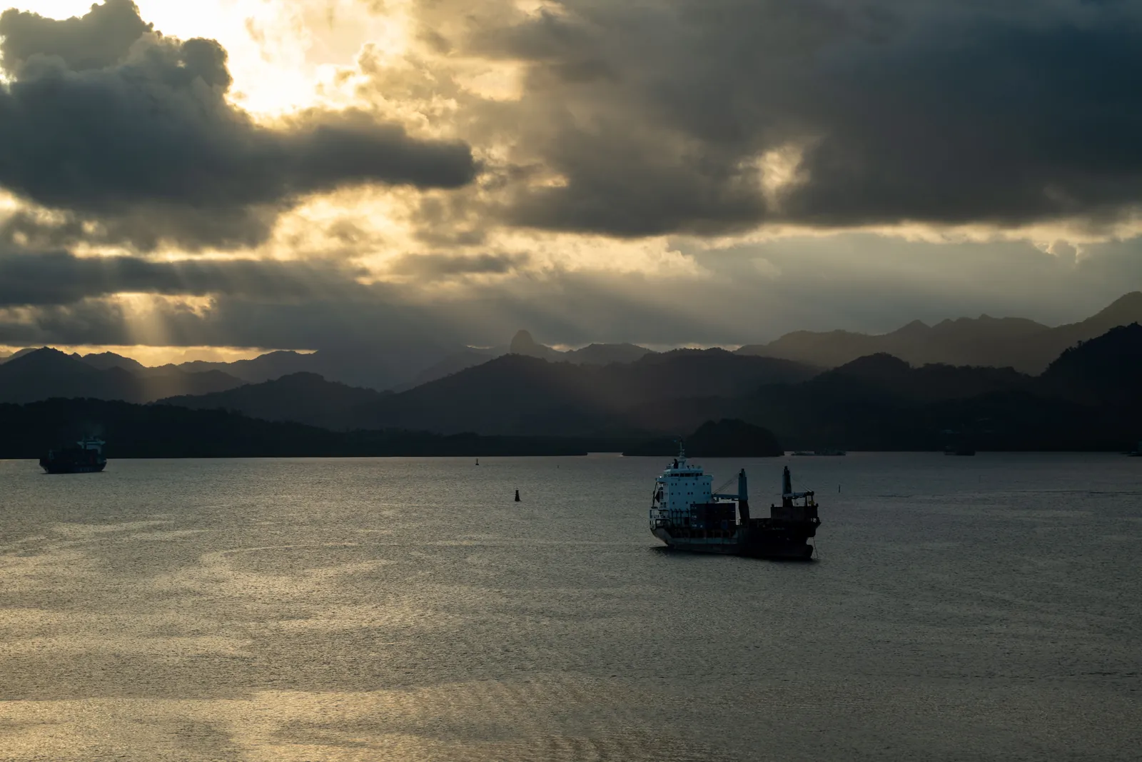 Cargo ships at anchor in a Fiji harbour at sunset, sunbeams breaking through heavy cloud over dark mountains
