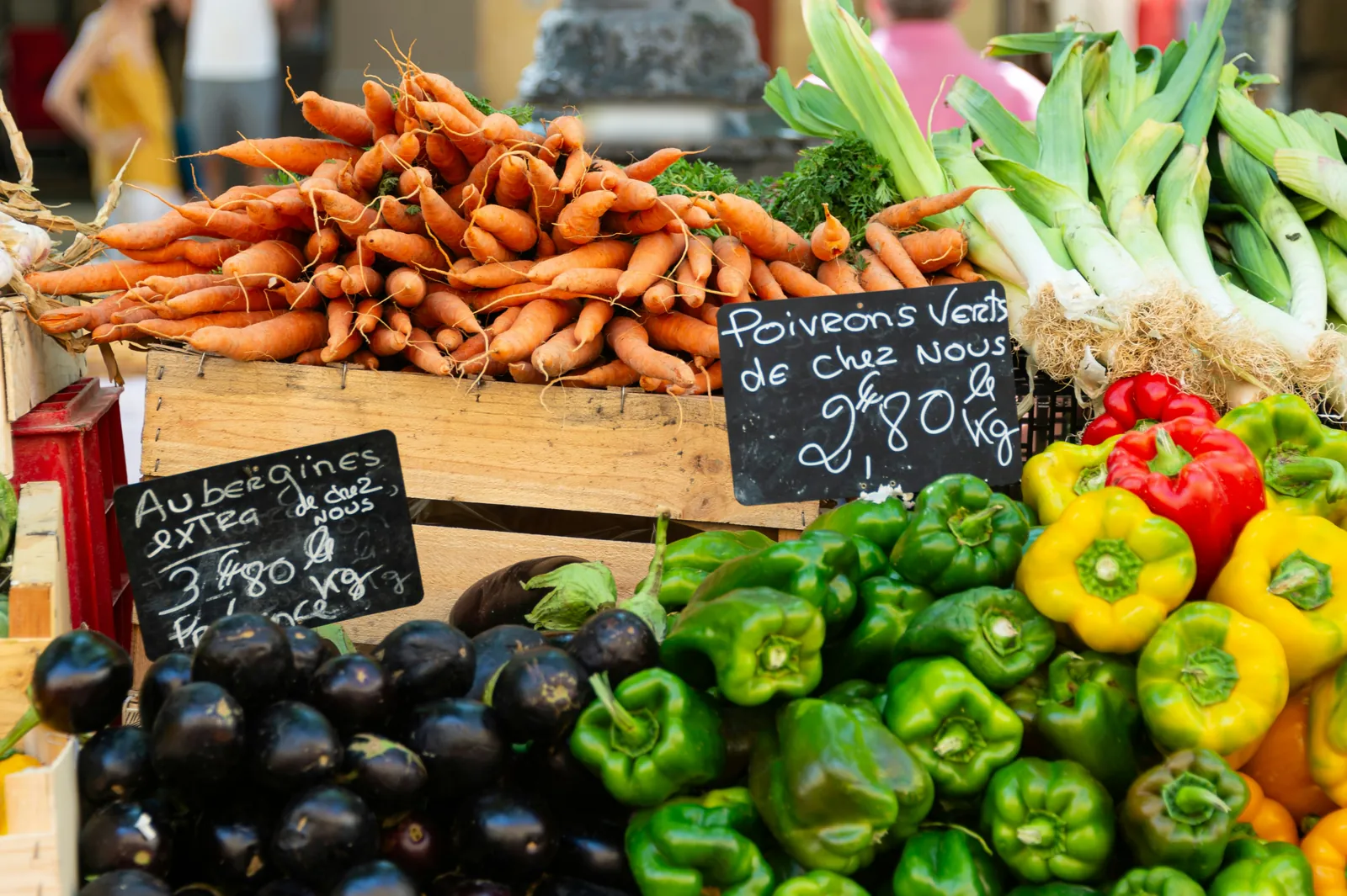 Market stall piled with bunches of carrots, leeks, glossy aubergines and red, yellow and green peppers, with handwritten chalkboard price signs in French