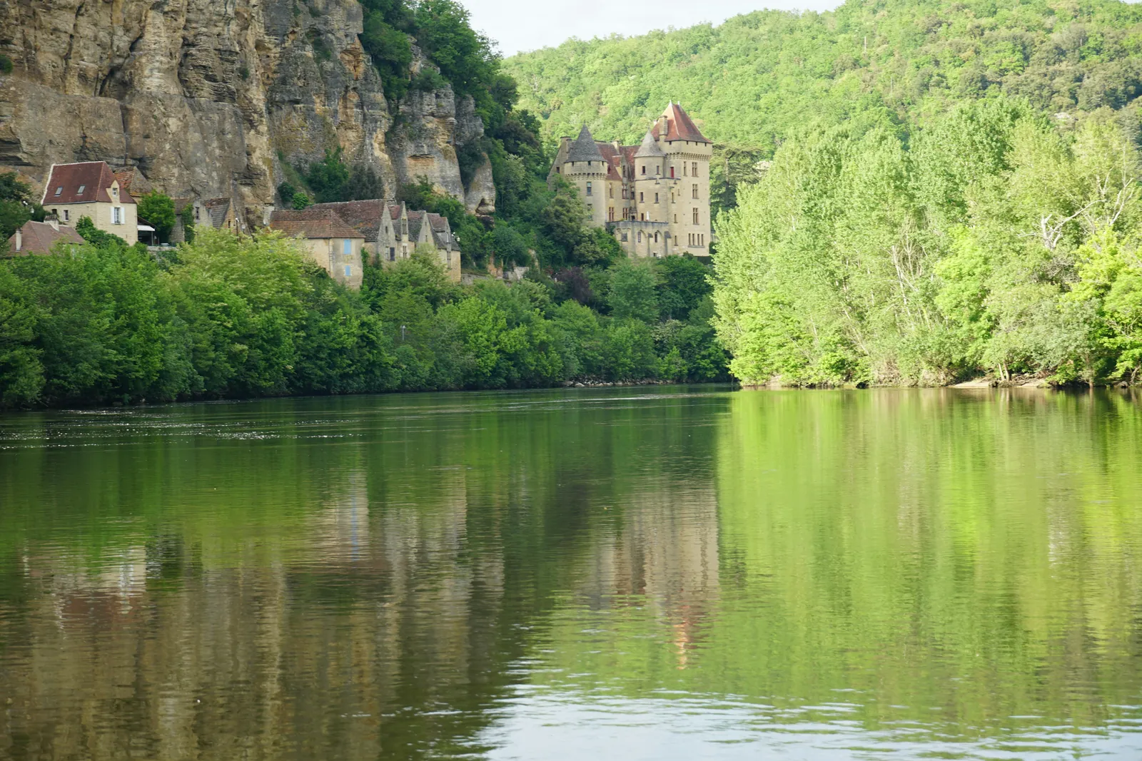 Golden stone houses and a turreted chateau tucked under a tall limestone cliff, with thick green trees reflected in a wide, still river