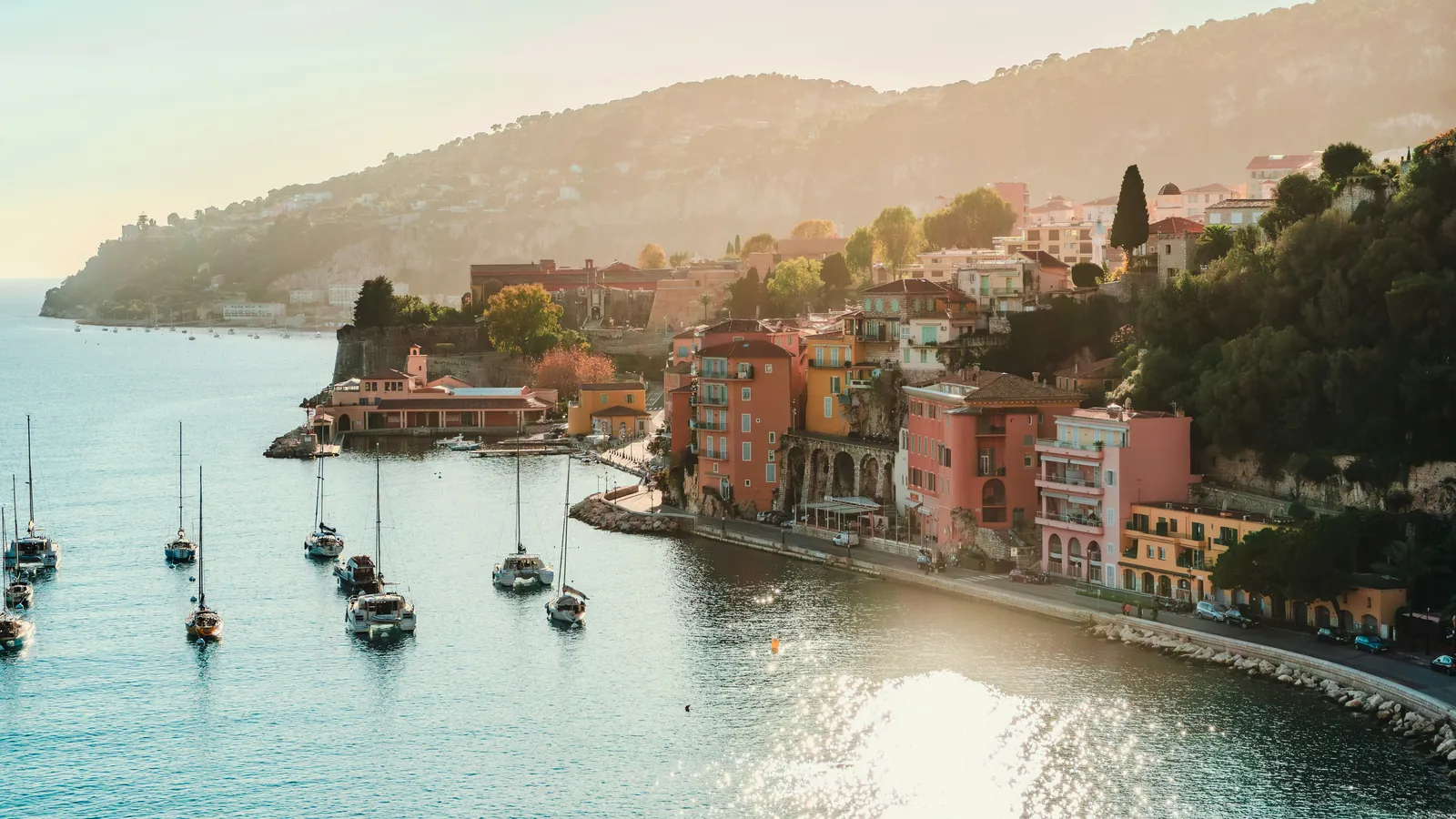 Pink and ochre old houses stacked along a curving waterfront road, with sailing boats moored in a calm bay and a wooded headland behind in soft morning light