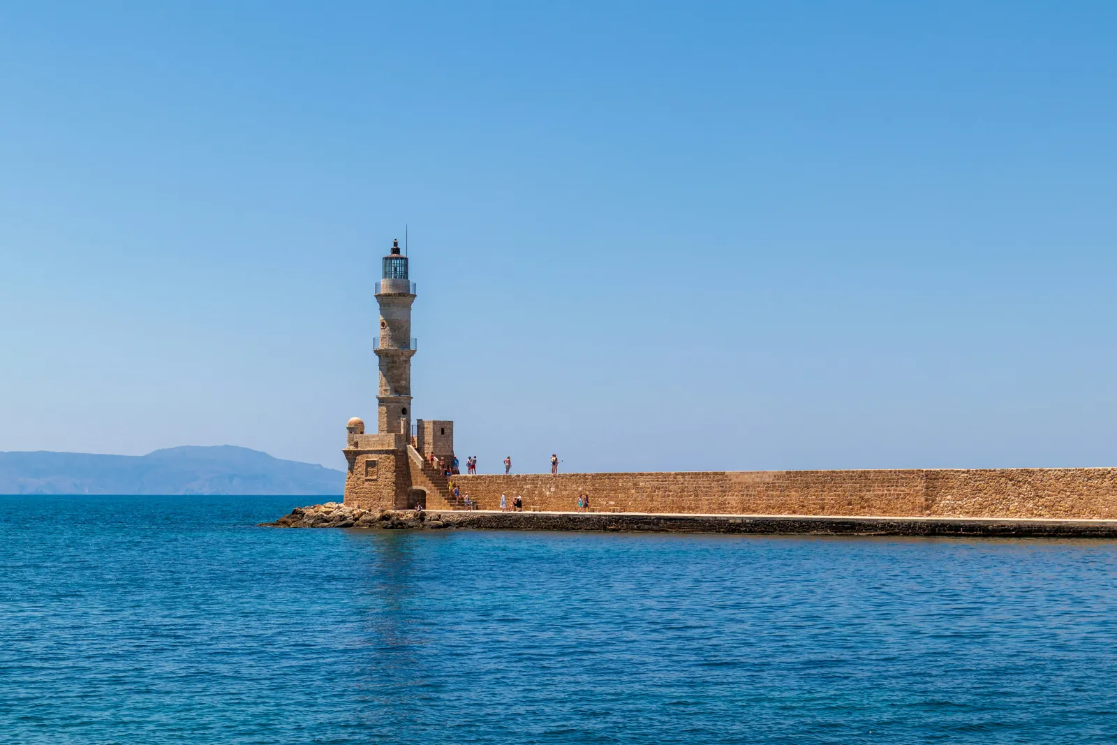 The stone lighthouse at the end of the old harbour breakwater in Chania, Crete, with calm blue sea and mountains on the horizon