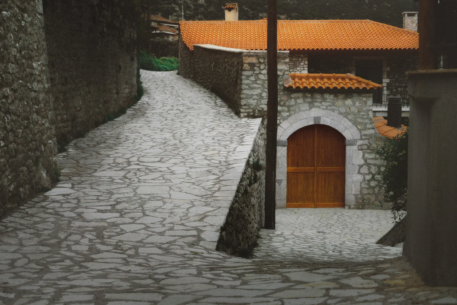 A stone-paved lane curving uphill past a stone house with an orange tiled roof and an arched wooden door, in the village of Dimitsana