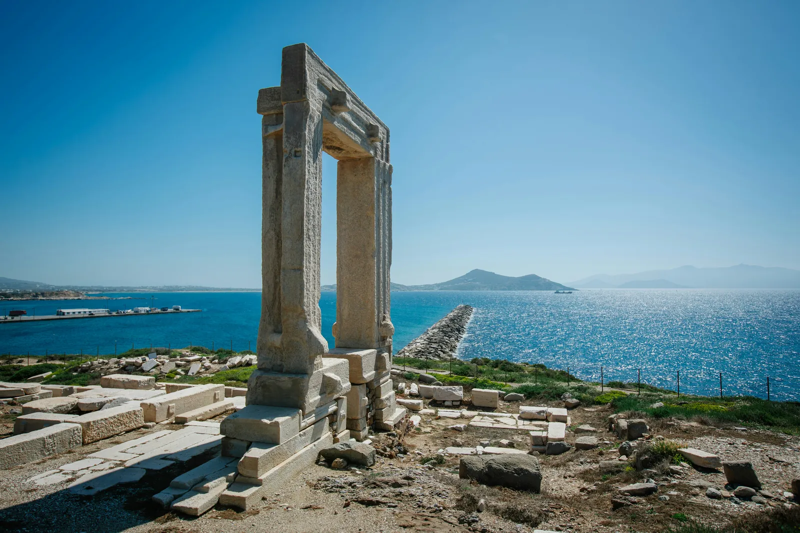 The marble Portara gateway of an ancient temple standing among fallen stones on a headland, with the blue Aegean and distant islands behind