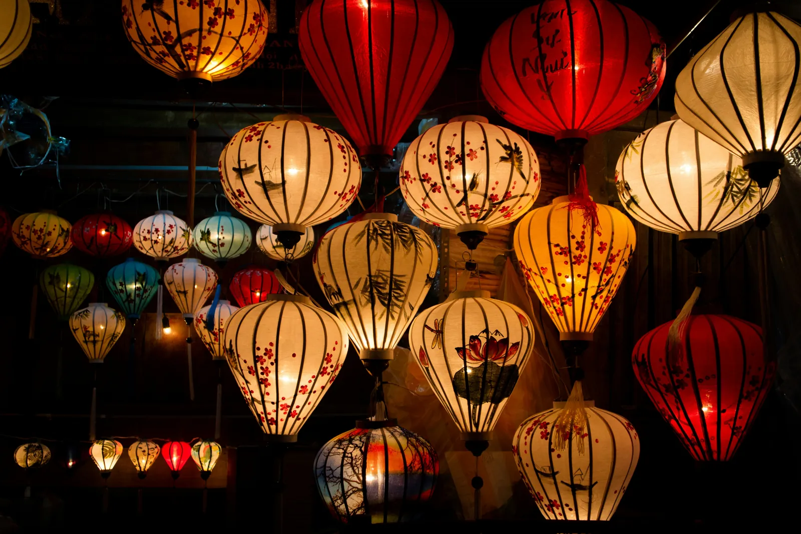 Silk lanterns in red, cream and orange, lit up and hanging close together against a dark background