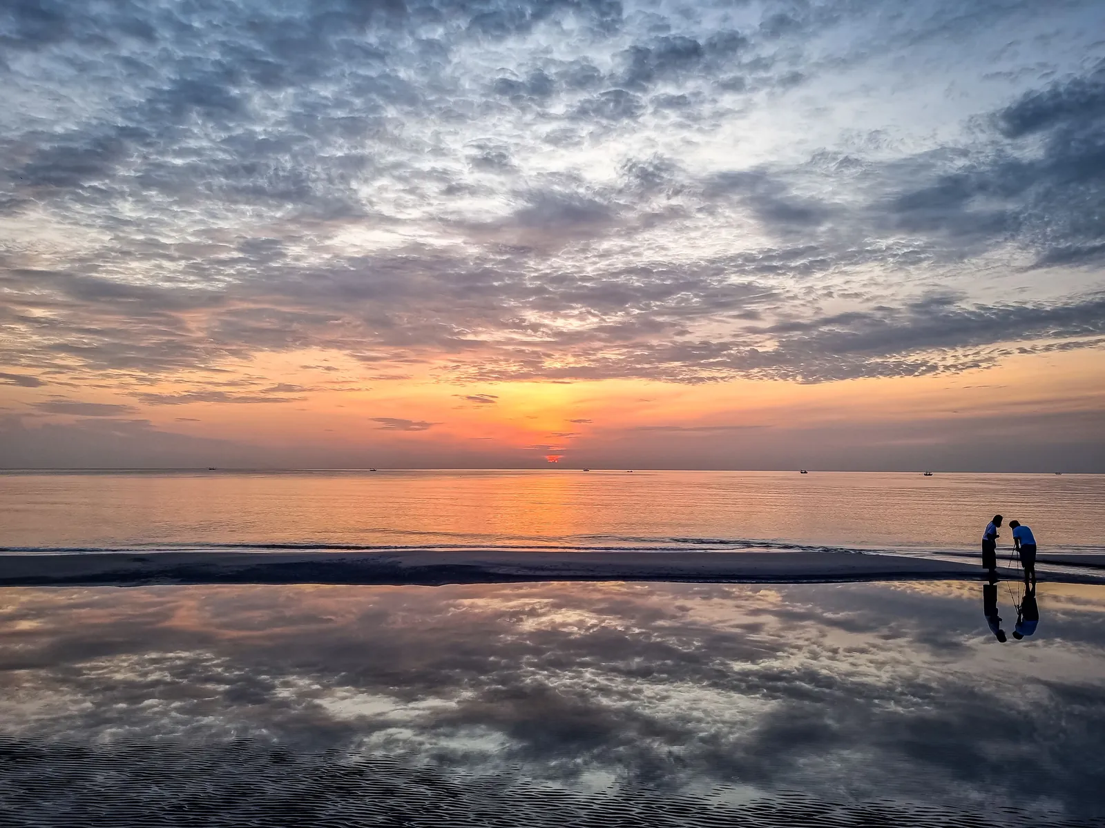 The sun low on a flat sea horizon under a sky of grey and orange dappled cloud, mirrored in a sheet of wet sand, with two small figures in silhouette at the water’s edge