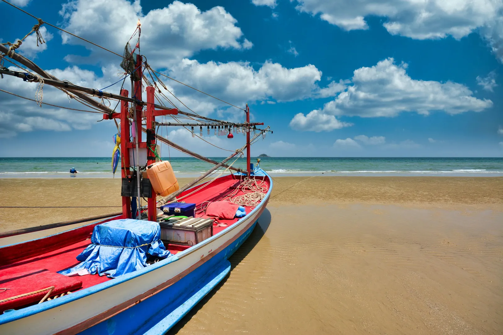 A wooden fishing boat with a red deck, blue and white hull and a frame of red posts hung with lamp bulbs, pulled up on flat wet sand, with a calm green sea, a small island on the horizon and white clouds in a blue sky