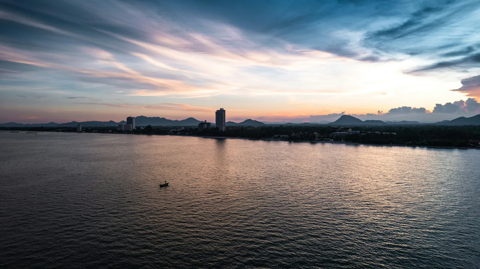 A calm sea reflecting a pink and blue sunset sky streaked with cloud, a single small boat in the middle, and a low dark coastline with a few tall buildings and hills along the horizon