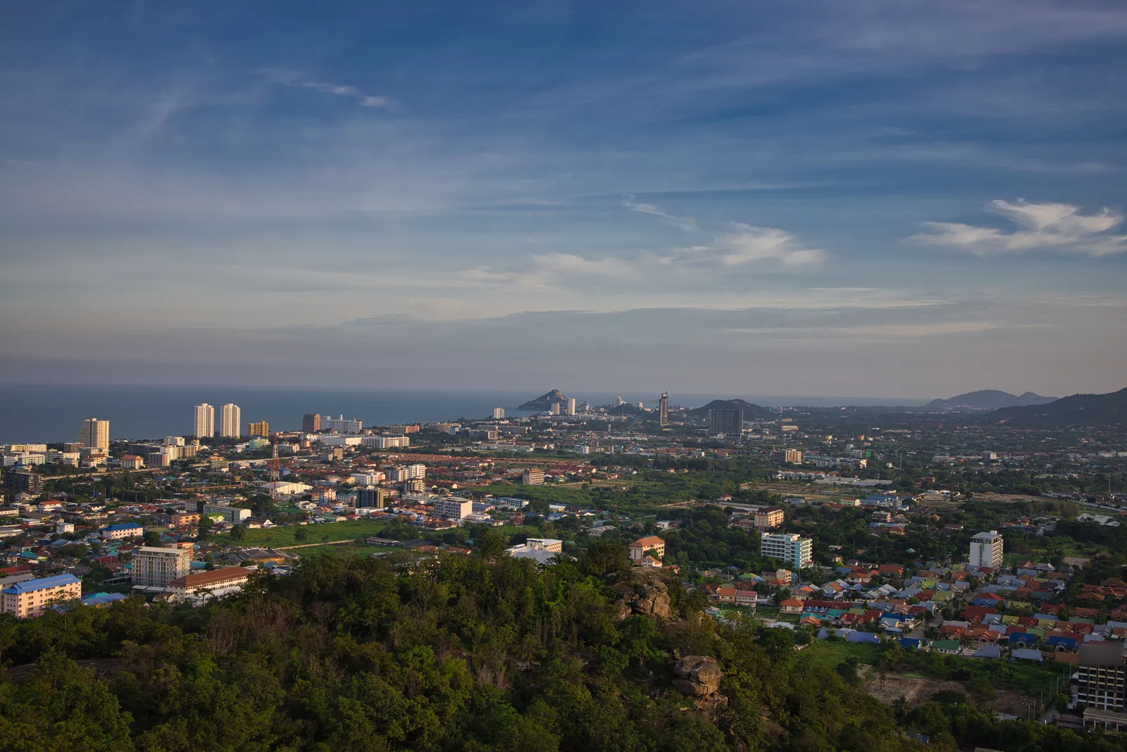 A town of low houses with coloured roofs, a few white tower blocks near the sea and green open ground, seen from a wooded hilltop in warm evening light, with the sea and pointed headlands on the horizon