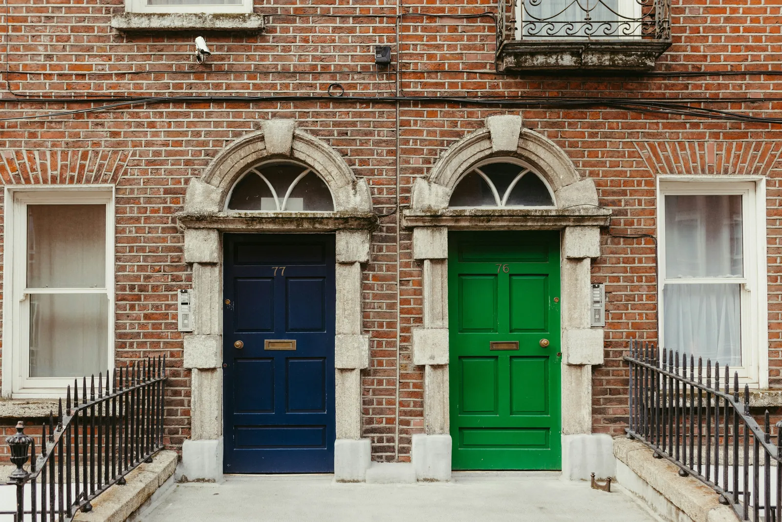 Two arched doorways side by side in a red brick terrace, one navy blue door and one bright green door, each with a fanlight above and black railings in front