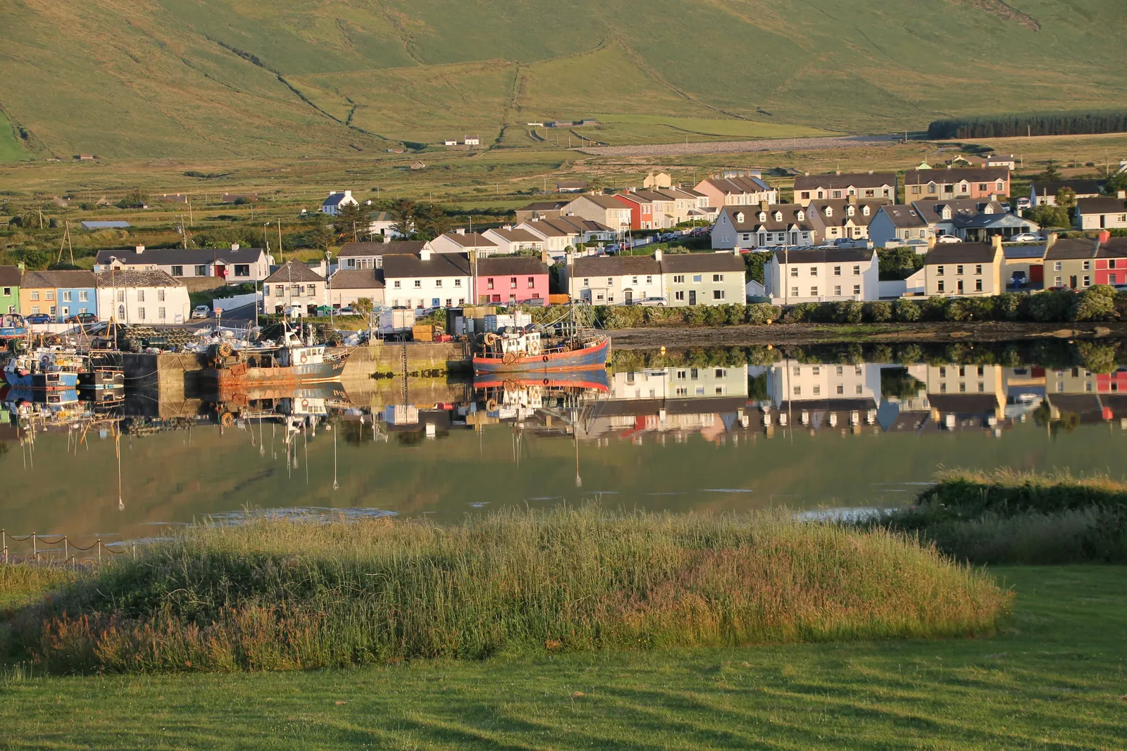 A row of white, pink and pastel houses along a still harbour with fishing boats tied up, the houses reflected in the water and a green hillside behind
