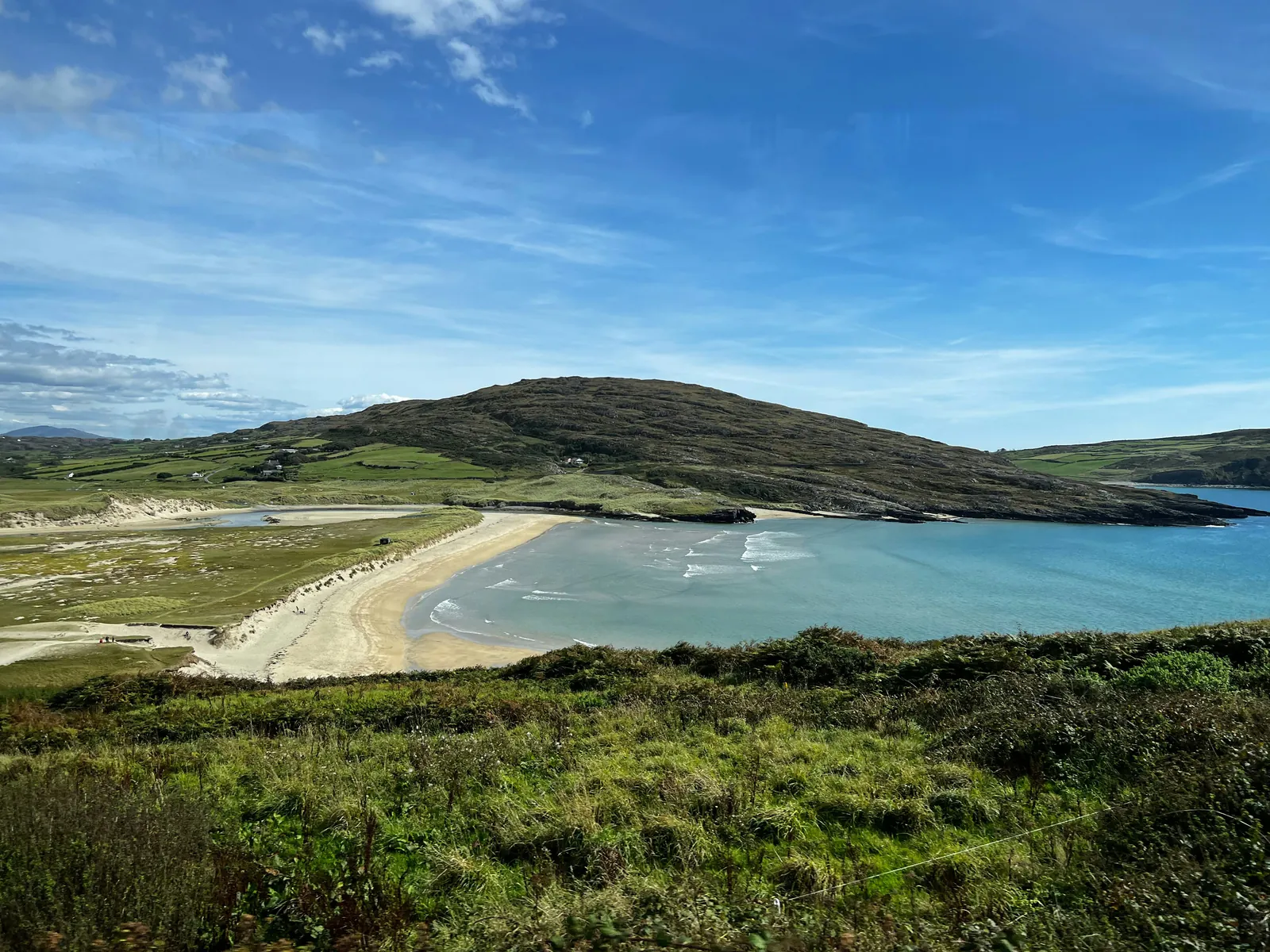 A wide sandy bay with turquoise water and small surf, backed by dunes and a green and brown hill with scattered farmhouses, seen from a grassy slope