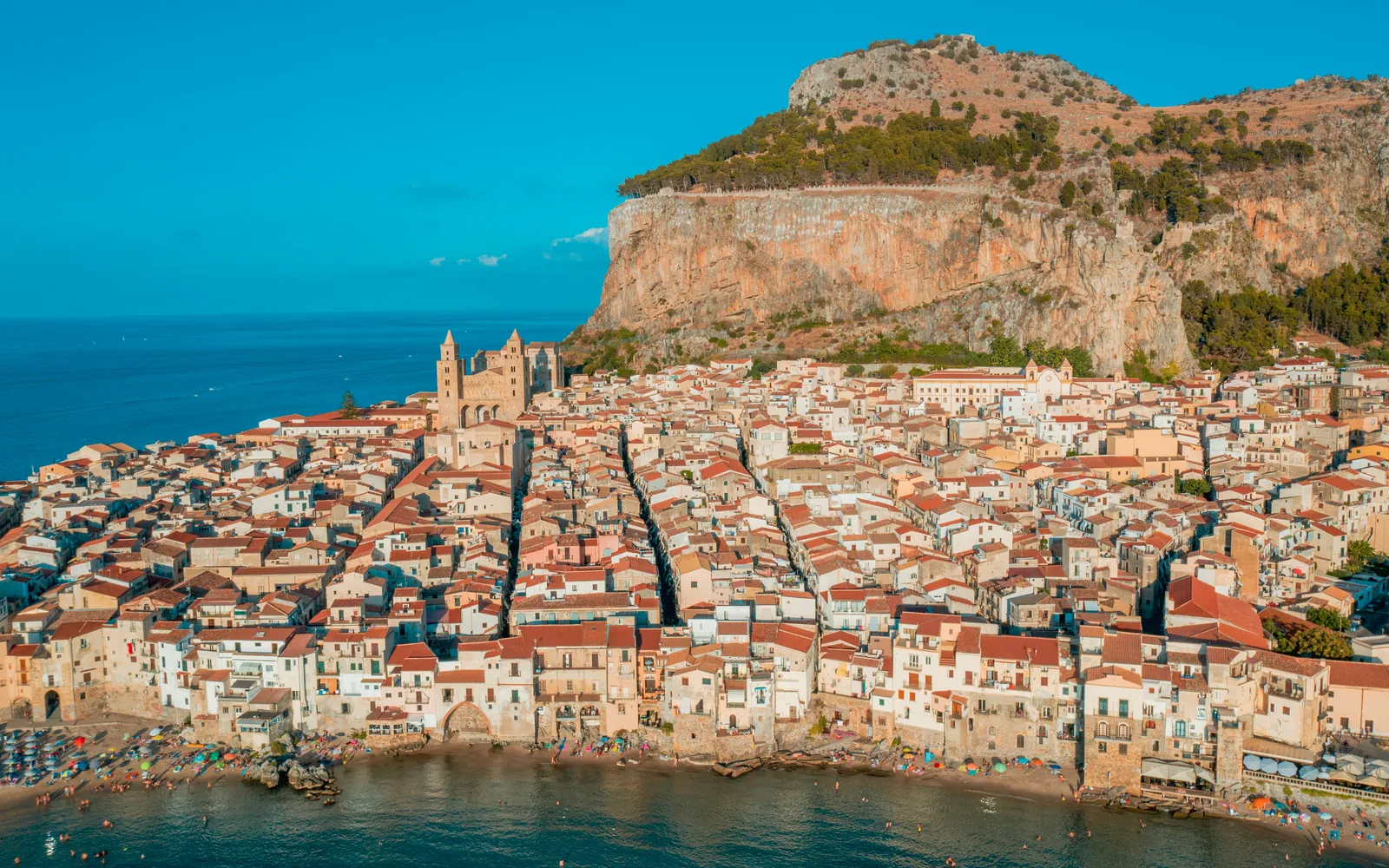 The old town of Cefalù packed along the shore below a great limestone headland, with the twin towers of its Norman cathedral and a small beach in front