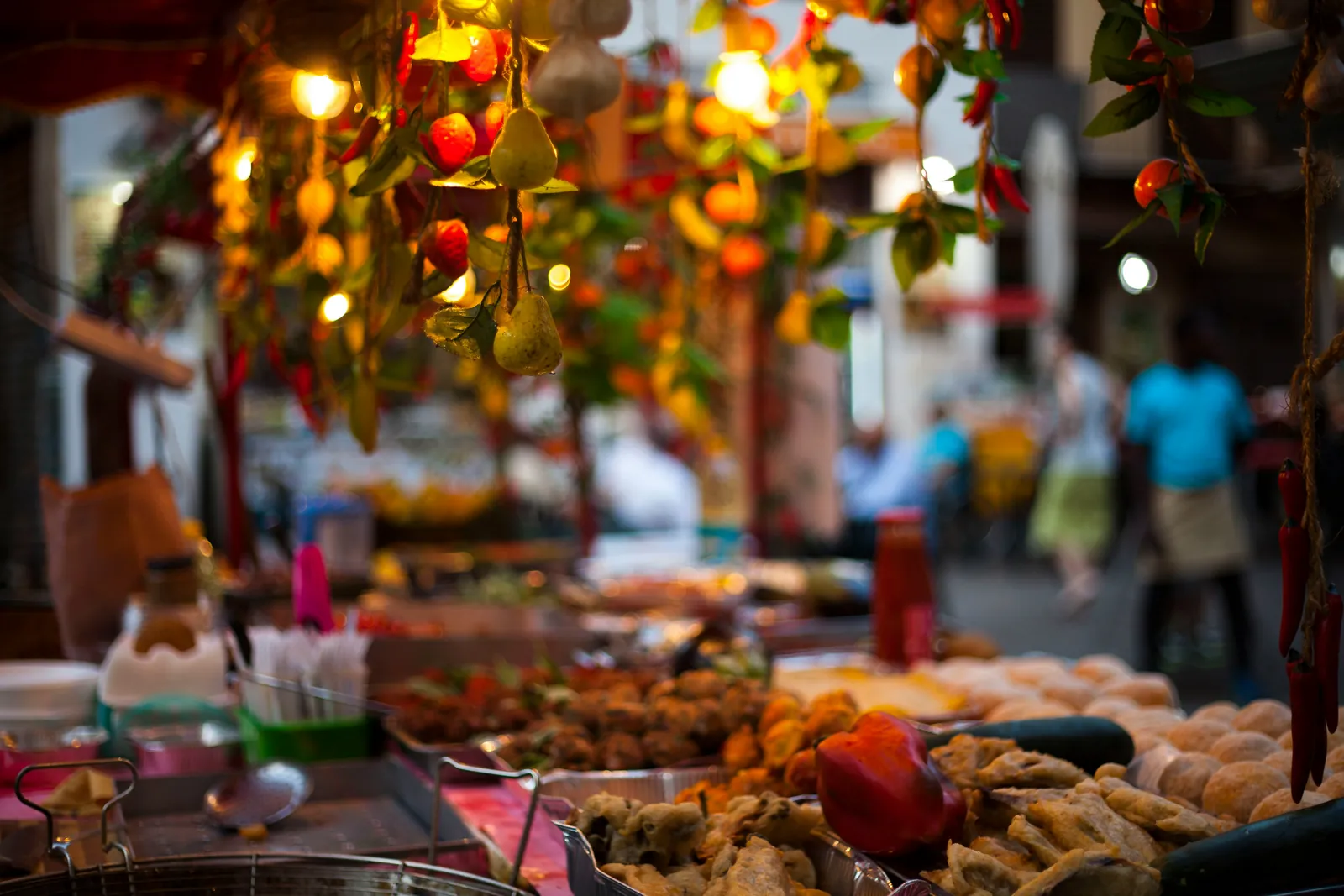 A market stall in Palermo with trays of fried vegetables and fritters under hanging strings of decorative fruit and warm lights, shoppers blurred behind