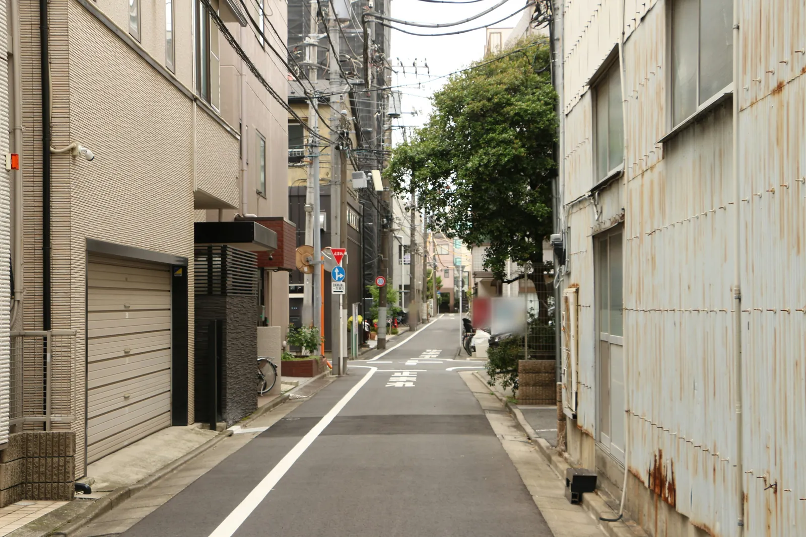 A narrow quiet Tokyo backstreet between plain apartment buildings and a rusty corrugated wall, with overhead power lines, a garage door, a tree and white road markings