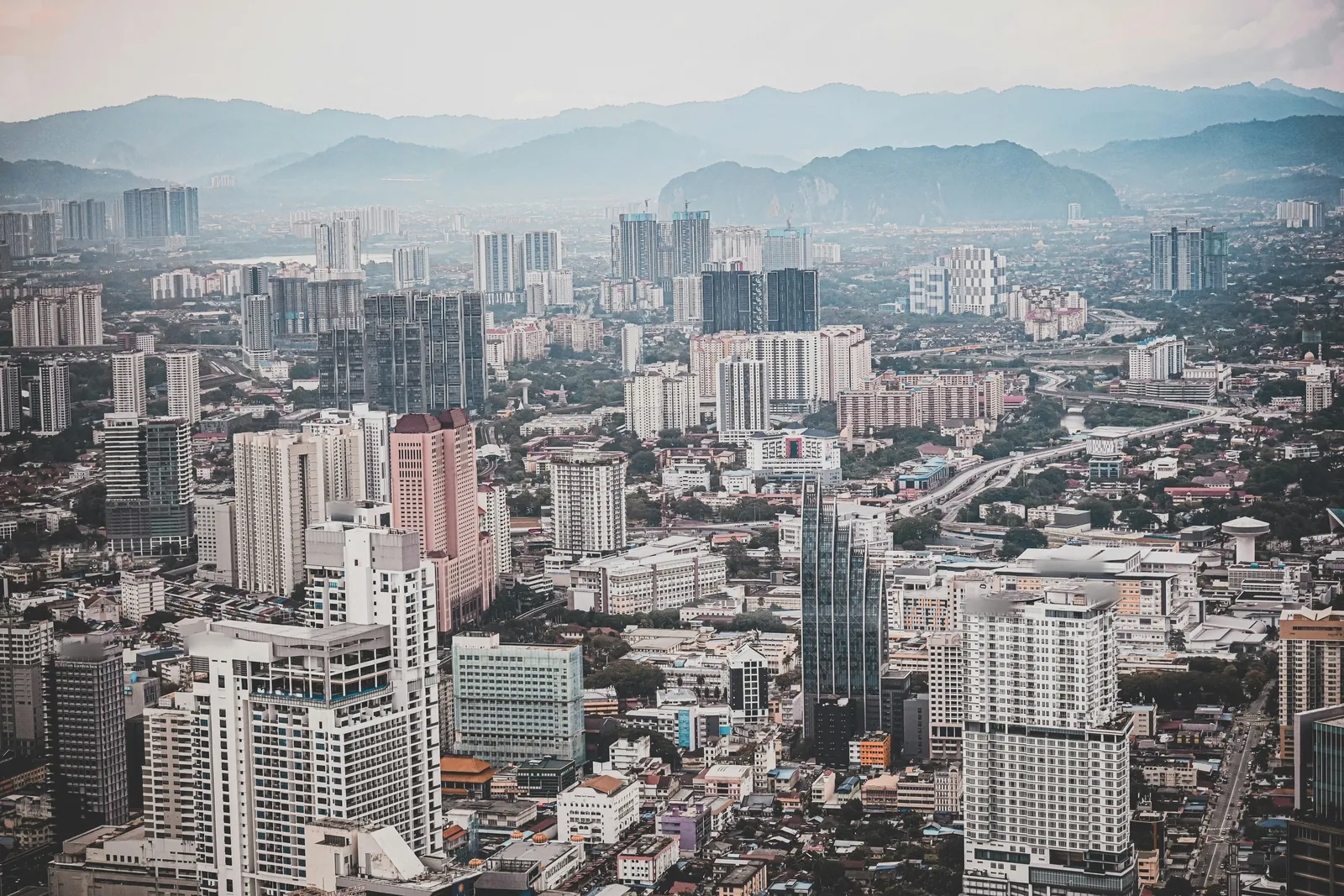 A dense spread of apartment towers, office blocks and low buildings seen from high above, with an elevated highway curving through, fading into haze towards rows of blue hills and a rocky outcrop
