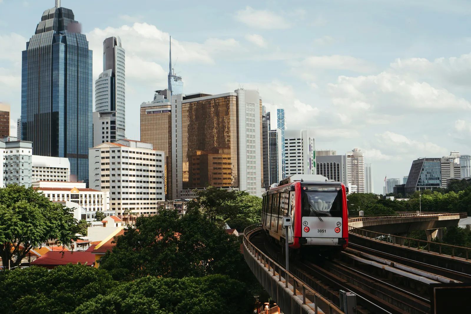 A red and white train on an elevated track curving round beside trees, with a cluster of glass and concrete office towers behind, one tall dark tower at the left and a thin spire in the distance, under a blue sky with cloud